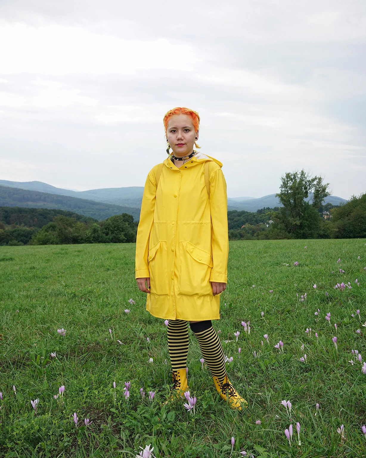 A young woman with orange hair wearing a yellow raincoat, black and yellow striped leggings, and yellow boots, standing in a green field with purple flowers, mountains in the background, and an overcast sky.