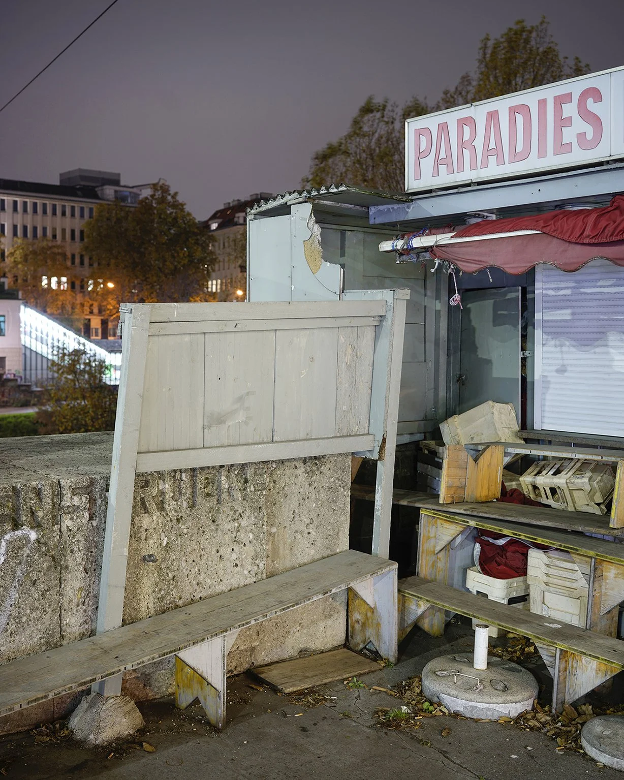 An abandoned food stand with a sign that reads 'PARADIES' in a city at night, showing worn-out wood and debris.