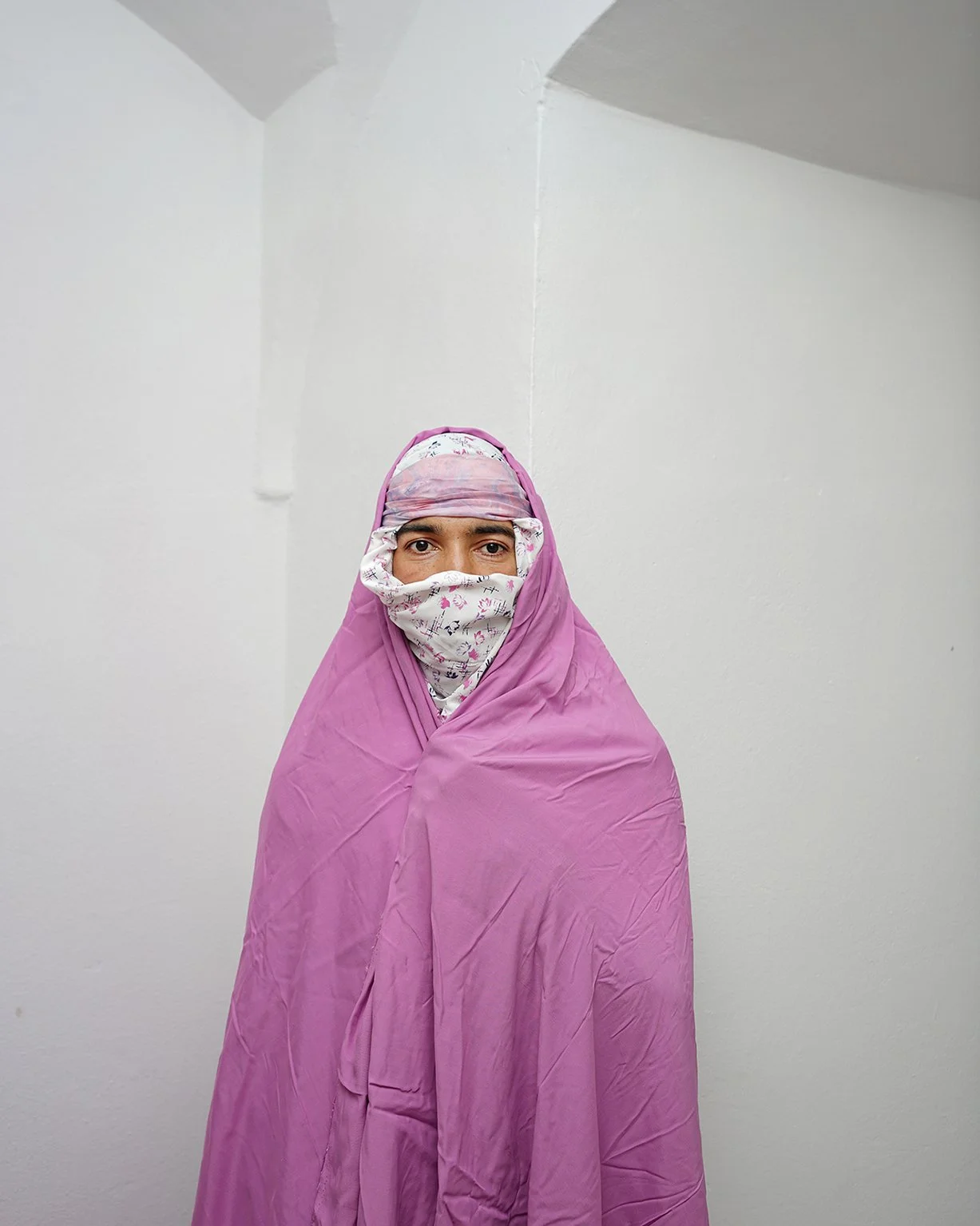 Woman wearing a pink shawl and a floral headscarf covering her hair and face, standing against a plain white wall.