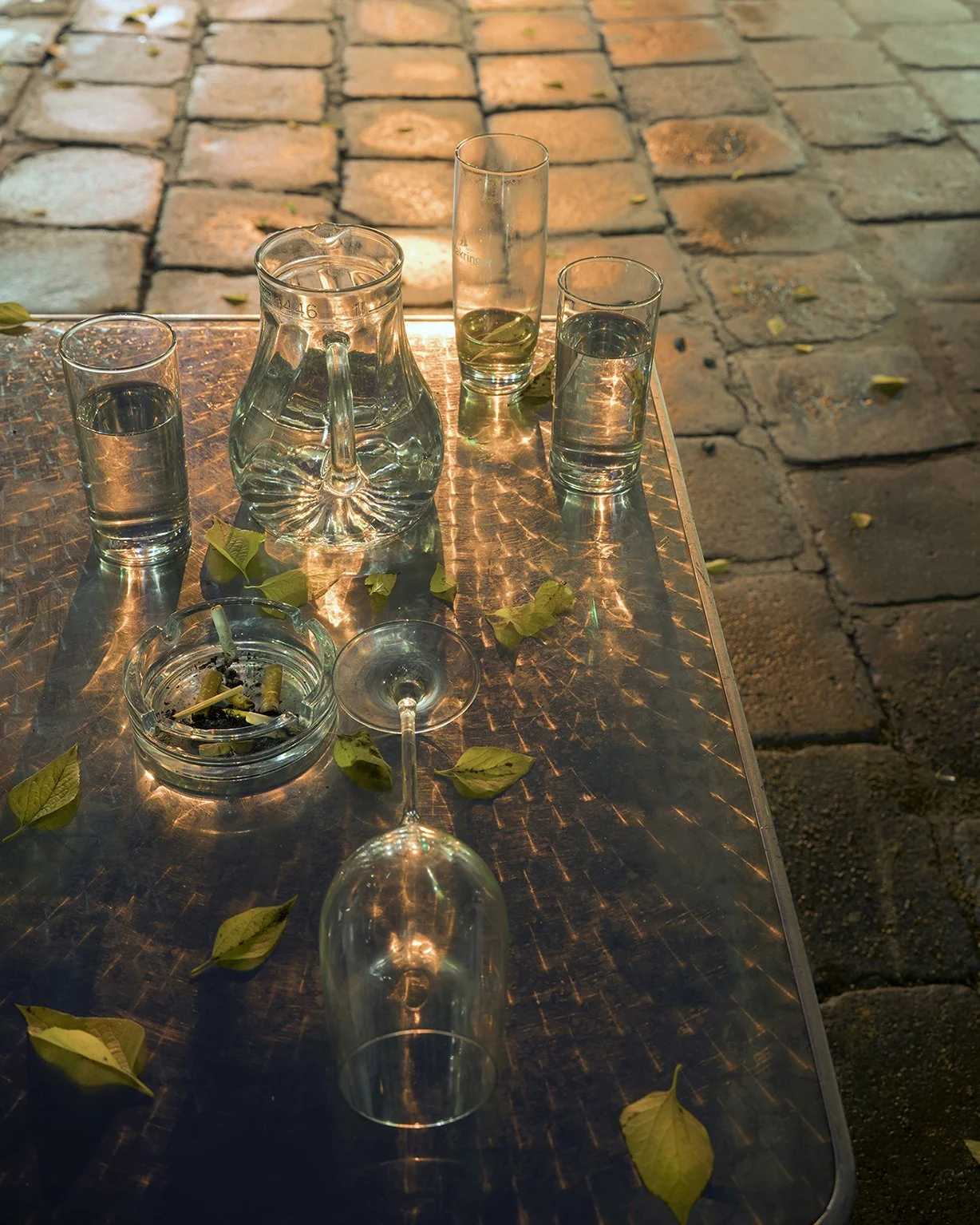 Empty glasses, a glass pitcher, a martini glass, a lit cigarette in an ashtray, and fallen leaves on a reflective metallic table outdoors.