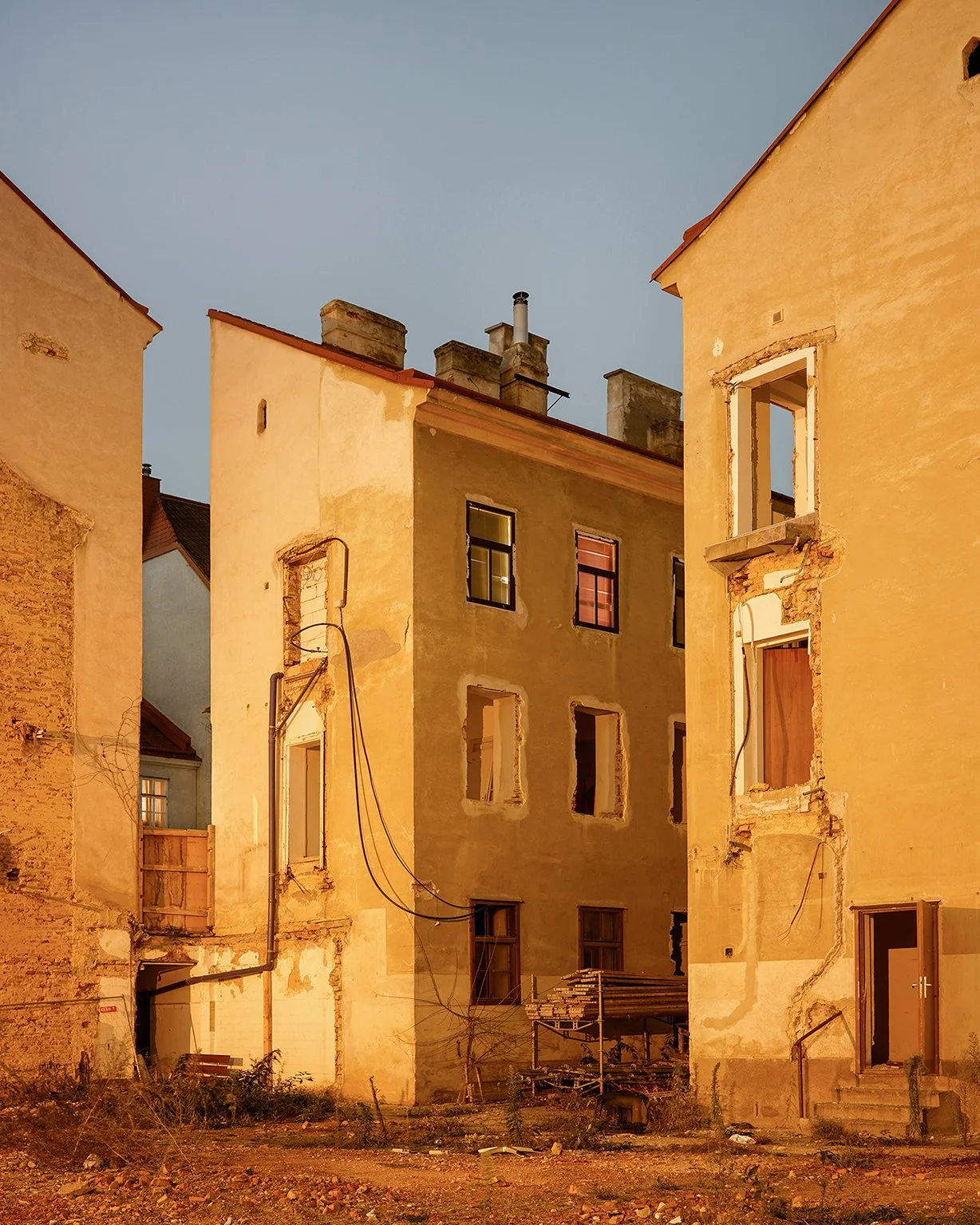 A group of old, weathered buildings with worn yellow and beige facades, some windows without glass and exposed brickwork, in a rustic setting during dusk.