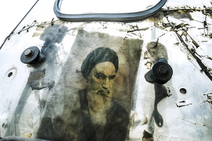 A worn, weathered photograph of a man with dark hair and a mustache, attached to a damaged, rusty surface with screws and some vines or wires crossing the top.