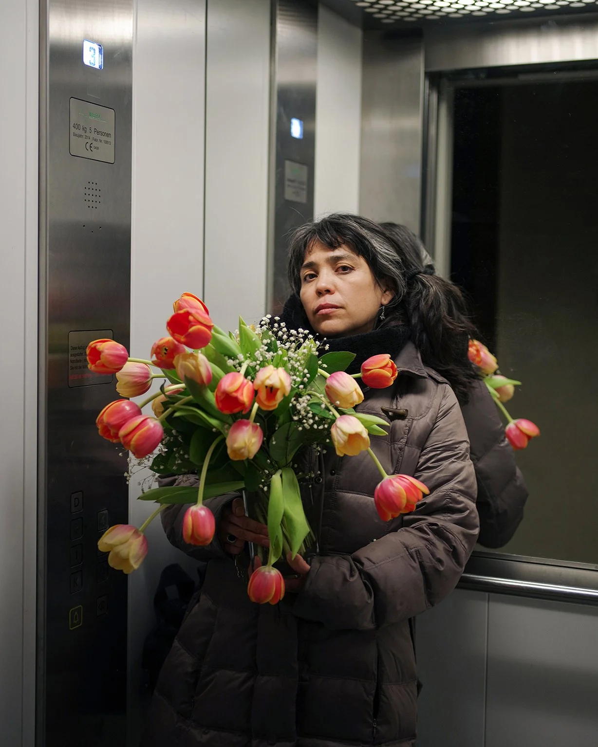 A woman with dark hair in a brown coat holding a large bouquet of pink and yellow tulips in an elevator.
