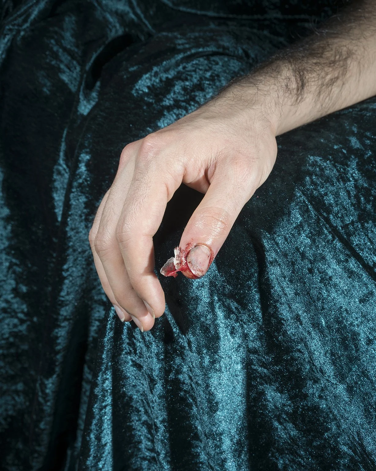 Close-up of a person's hand with a bloody finger, holding a piece of broken glass, against a blue velvet fabric background.