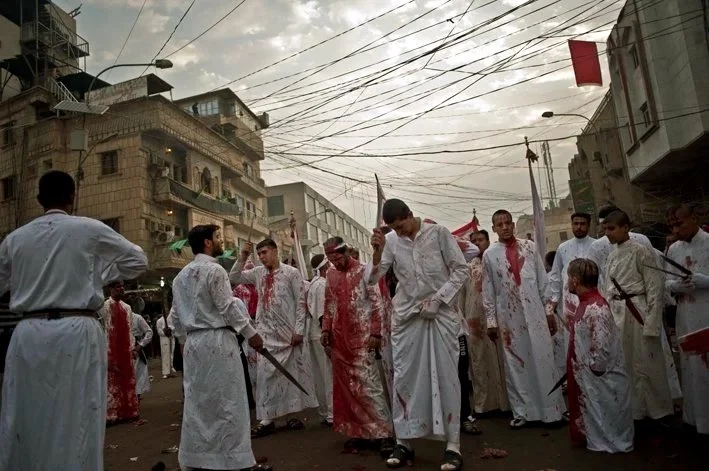 Group of men dressed in white traditional garments with some bloodstains, walking on a city street surrounded by buildings and overhead wires, participating in a religious or cultural procession.