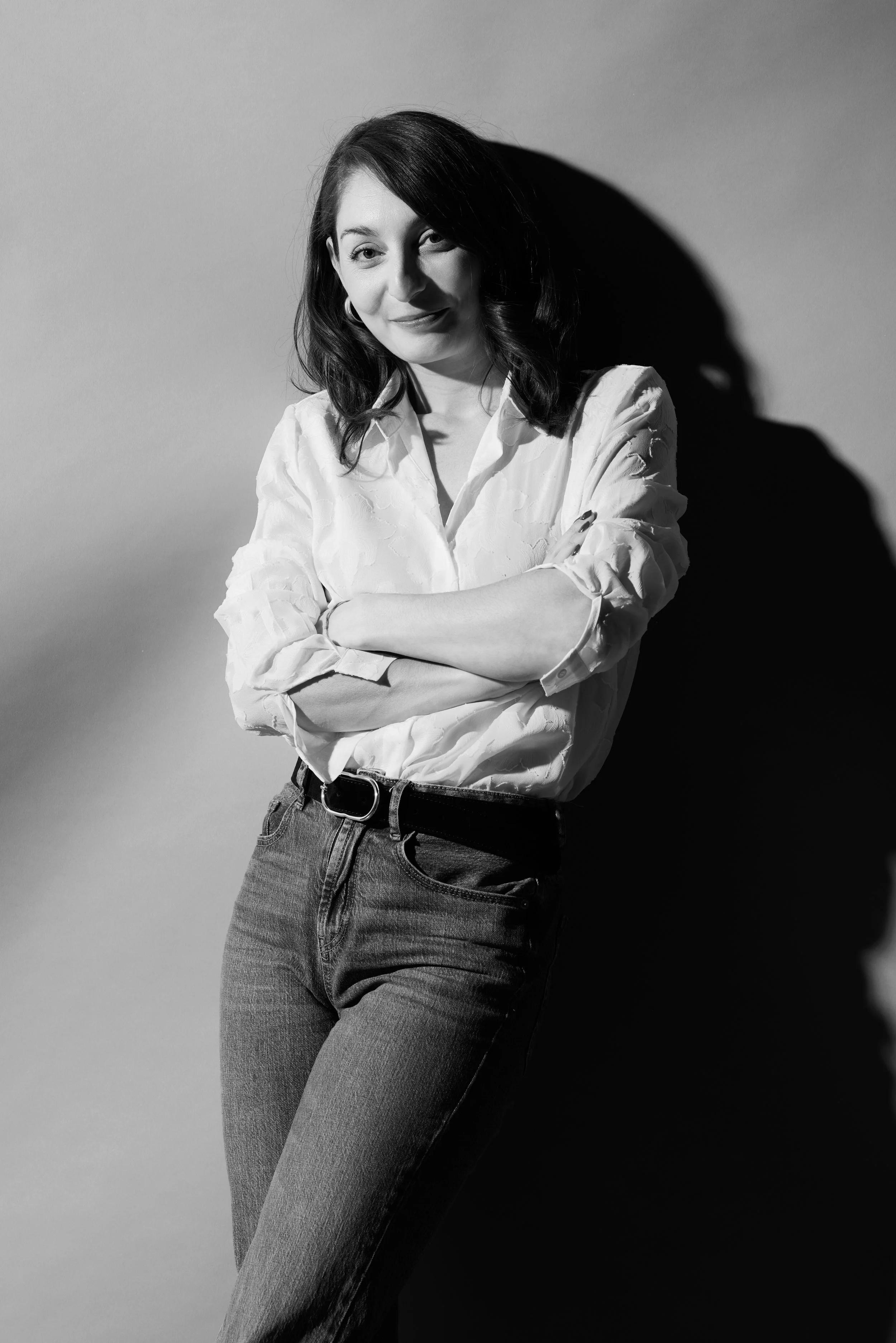 Jeune femme avec les bras croisés, portant une chemise blanche roulée aux manches, et un jean, se tenant contre un mur, en noir et blanc, avec un éclairage créant une ombre distinctive.