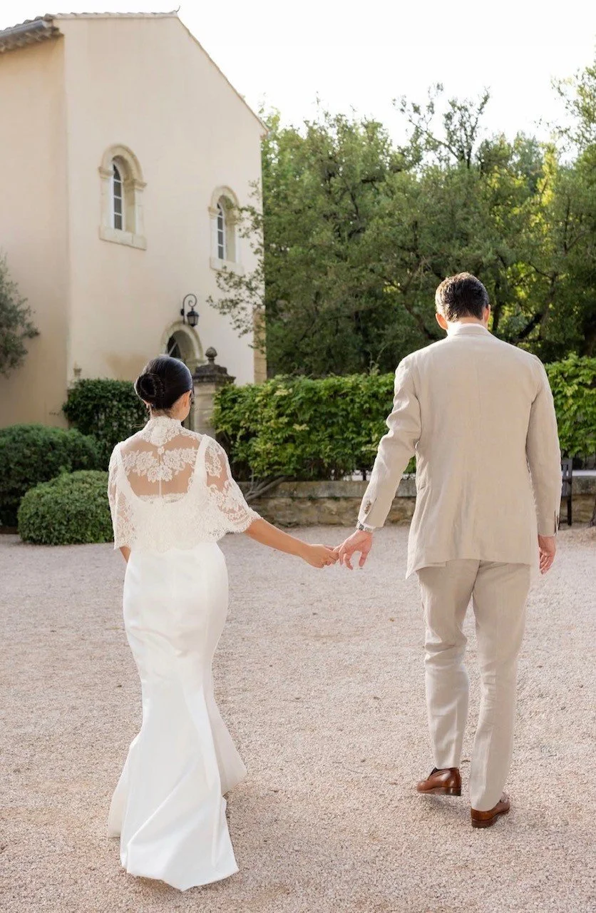 Un couple habillé en blanc marche dans un jardin, main dans la main, avec une maison en pierre à l'arrière-plan.
