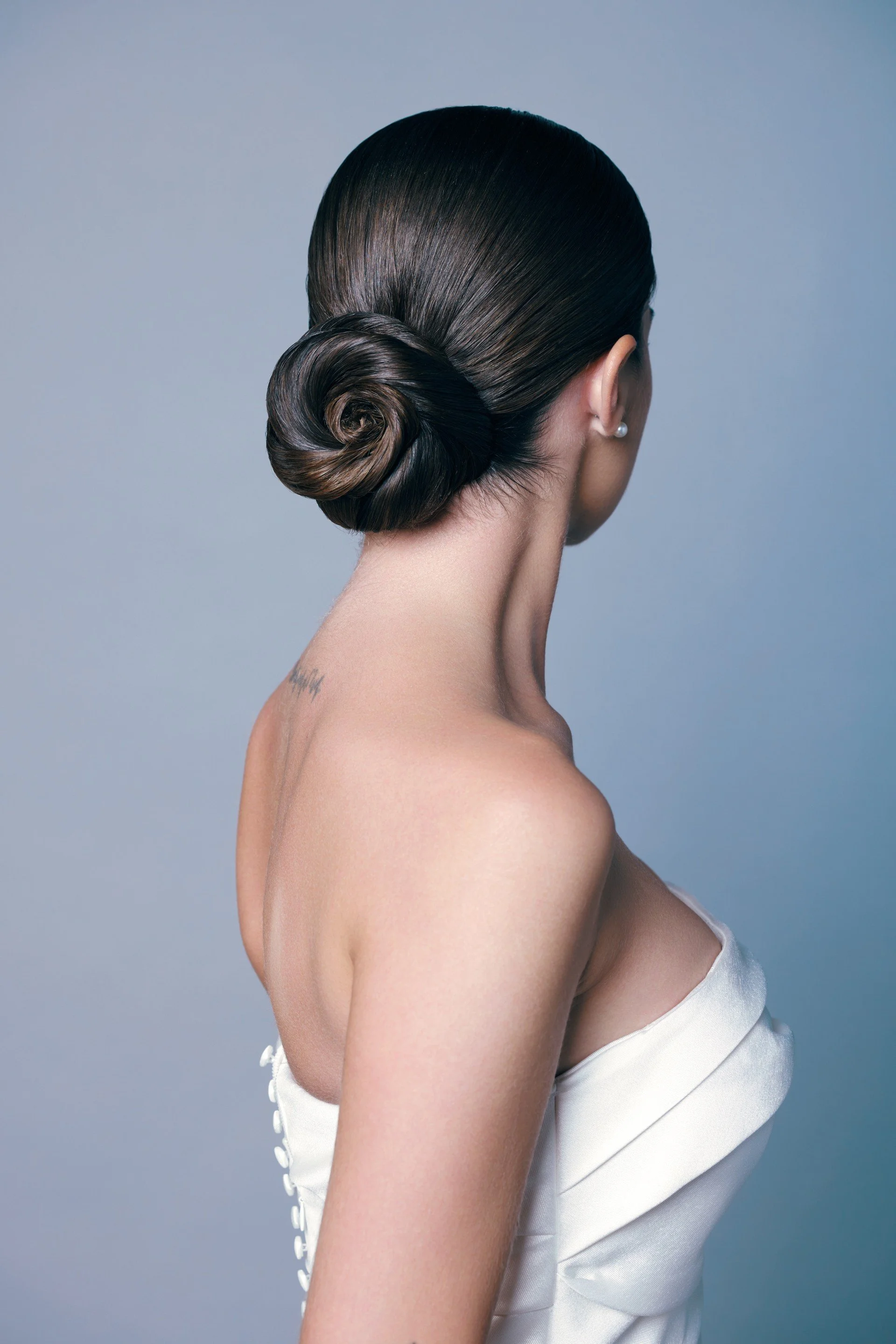 Une femme vue de dos, avec des cheveux coiffés en chignon, portant une perle à l'oreille et une robe de mariée