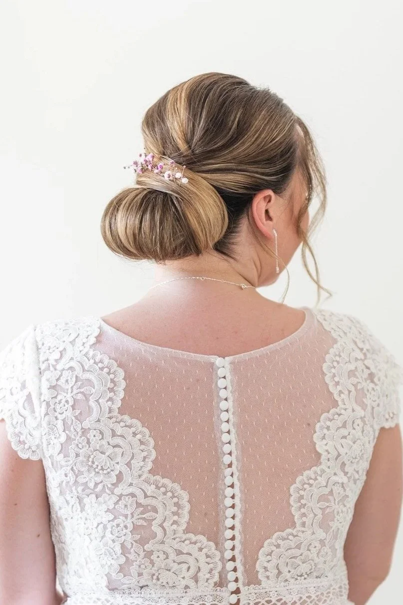 Une mariée dont la coiffure est un chignon avec des fleurs et portant une robe en dentelle vue de dos