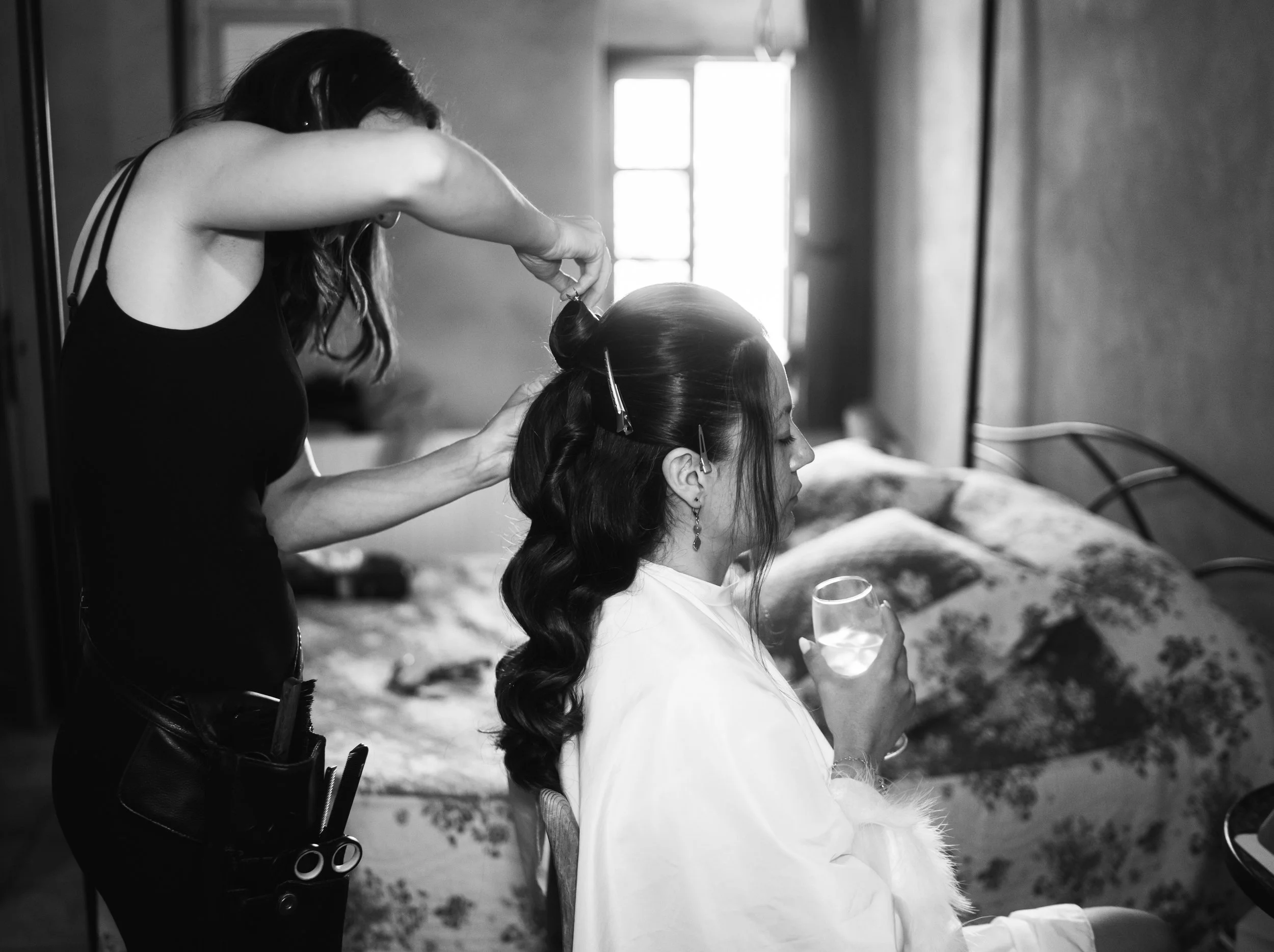 Une femme préparant les cheveux d'une autre femme qui porte une robe blanche et des boucles d'oreilles, en intérieur, en noir et blanc.