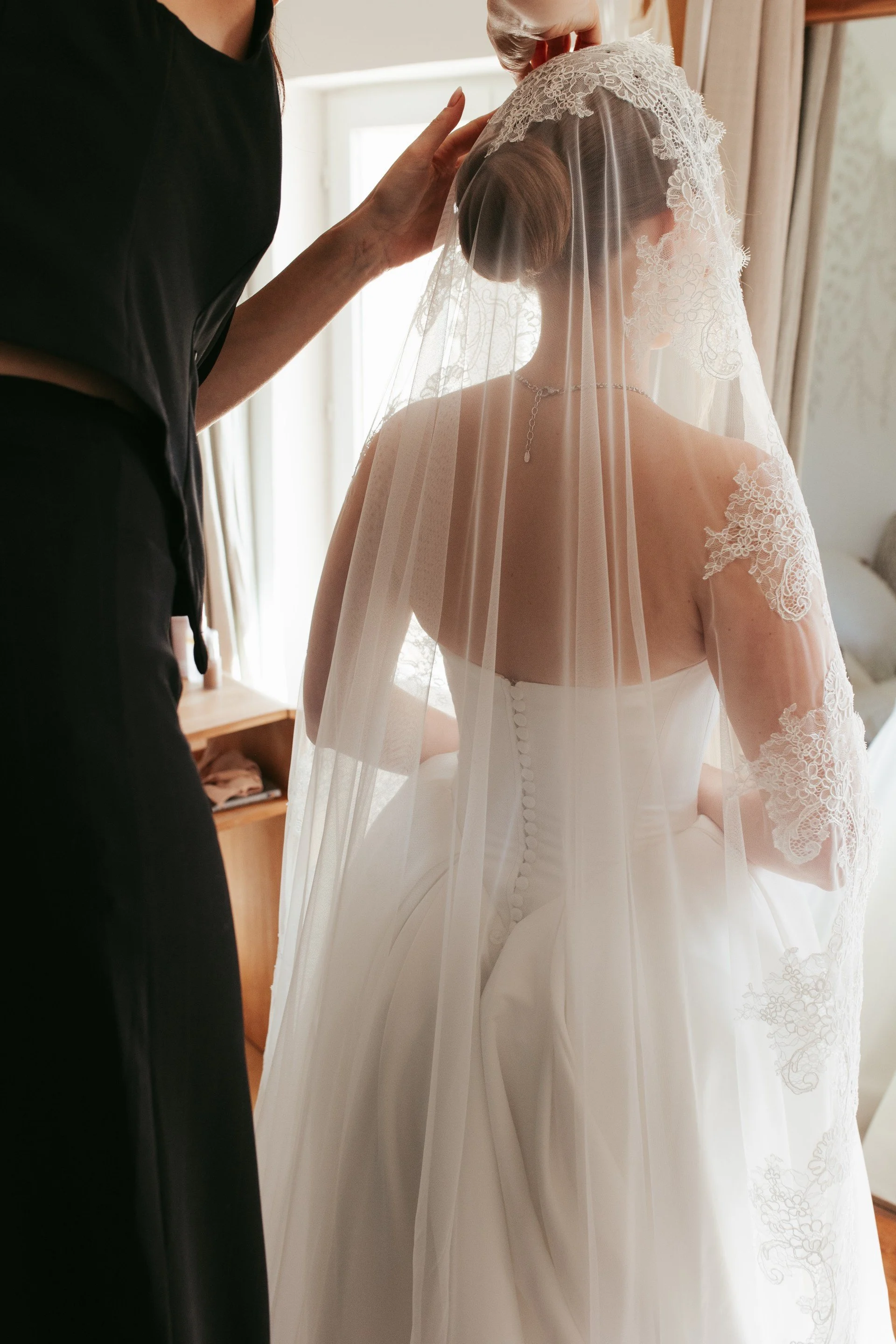 Femme en robe de mariage blanche portant un voile, vue de dos, devant une fenêtre, en train de se préparer.