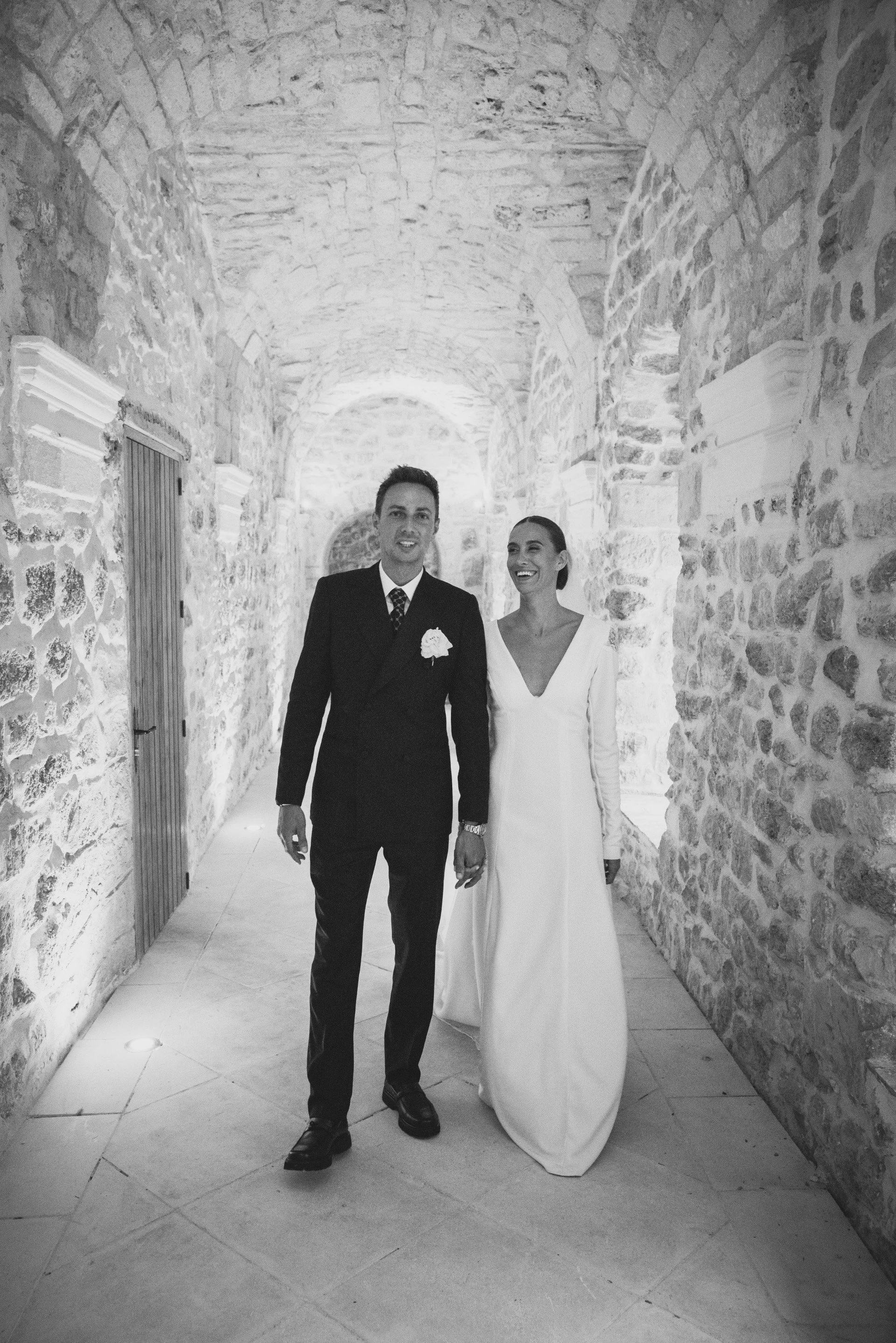 Un couple marié marche main dans la main dans un couloir en pierre, souriant. La mariée porte une robe blanche longue à manches longues et le marié un costume noir avec une boutonnière blanche.