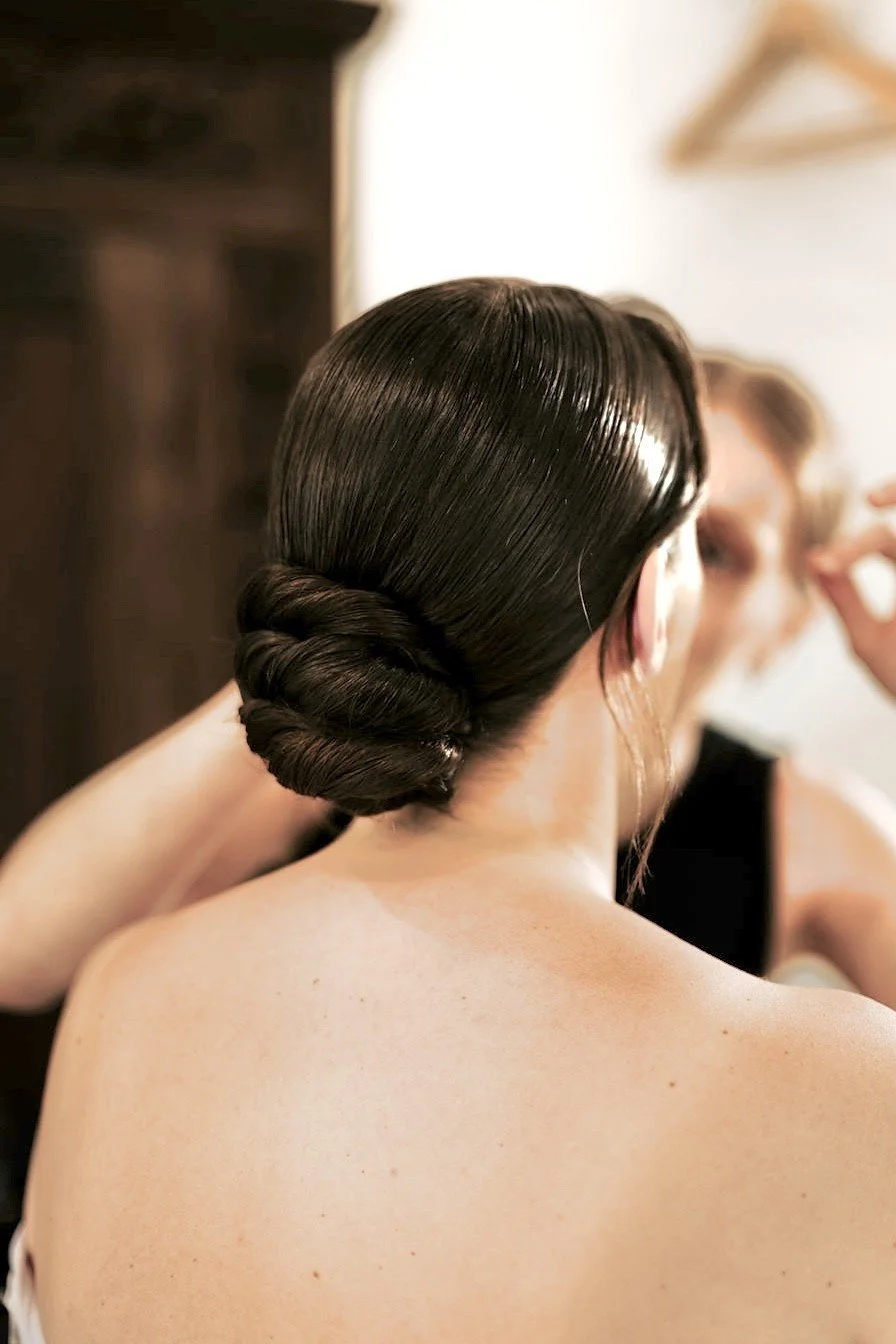 Une femme avec des cheveux noirs, coiffés en un chignon élégant, est en train de se faire maquiller par une autre personne, dans une chambre dans un domaine