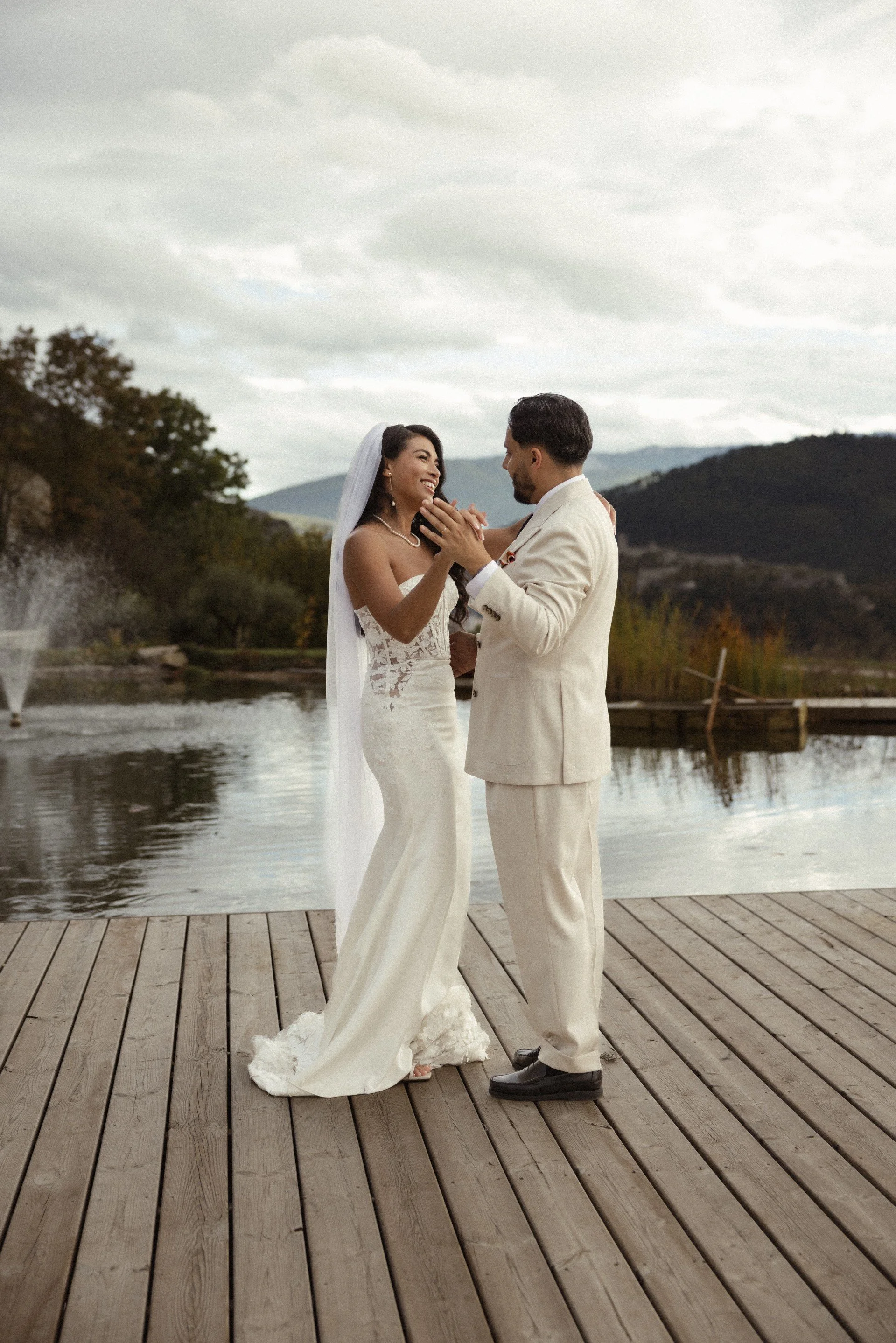 Un couple en mariage, se tenant la main en regardant dans les yeux, sur une plateforme en bois au bord de l'eau, avec un paysage de montagnes et de arbres en arrière-plan, par une journée nuageuse.