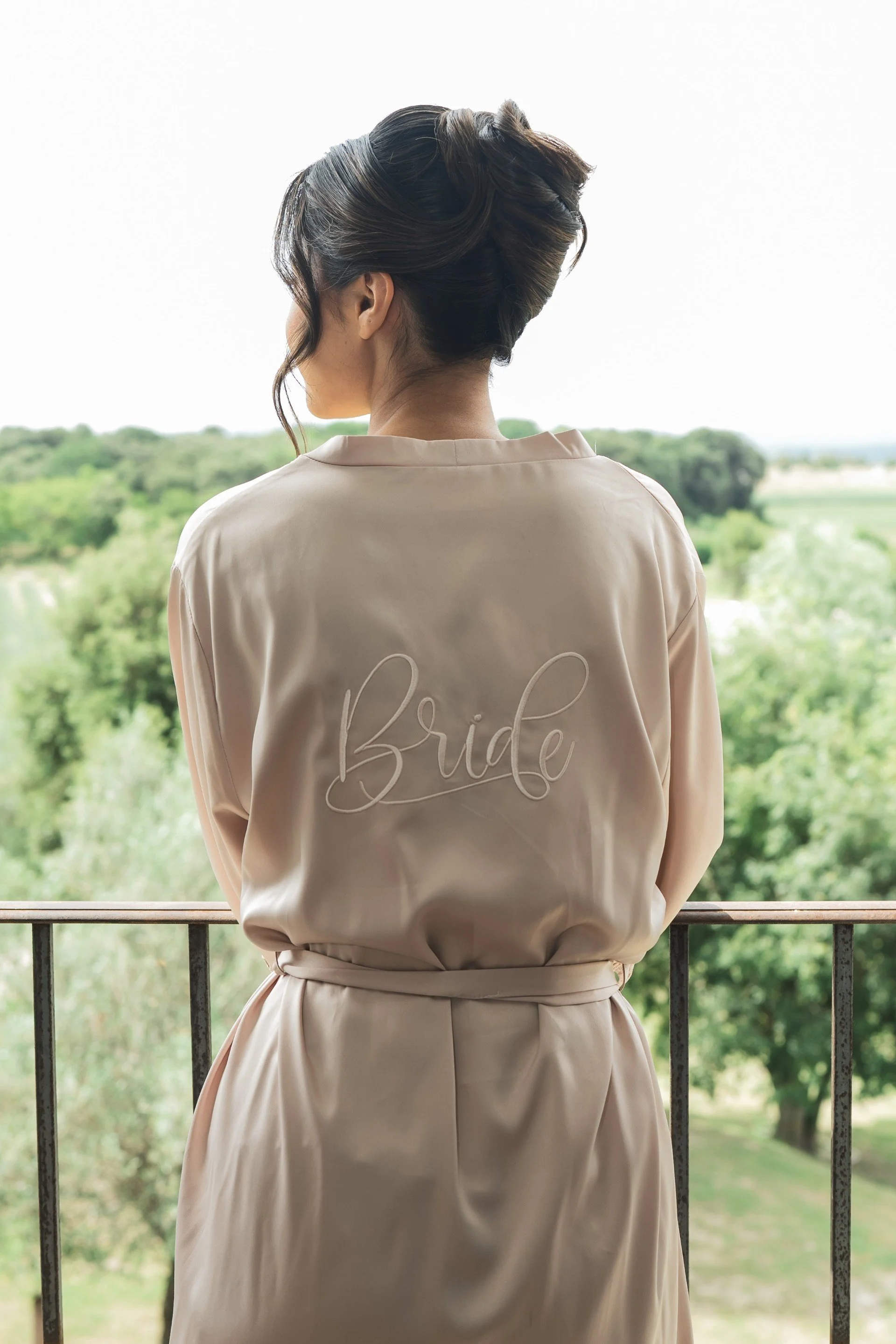 Une femme coiffée d'un chignon, vue de dos portant un peignoir de mariée rose avec l'inscription "Bride" brodée sur le dos, regardant un paysage verdoyant au-delà d'une balustrade.