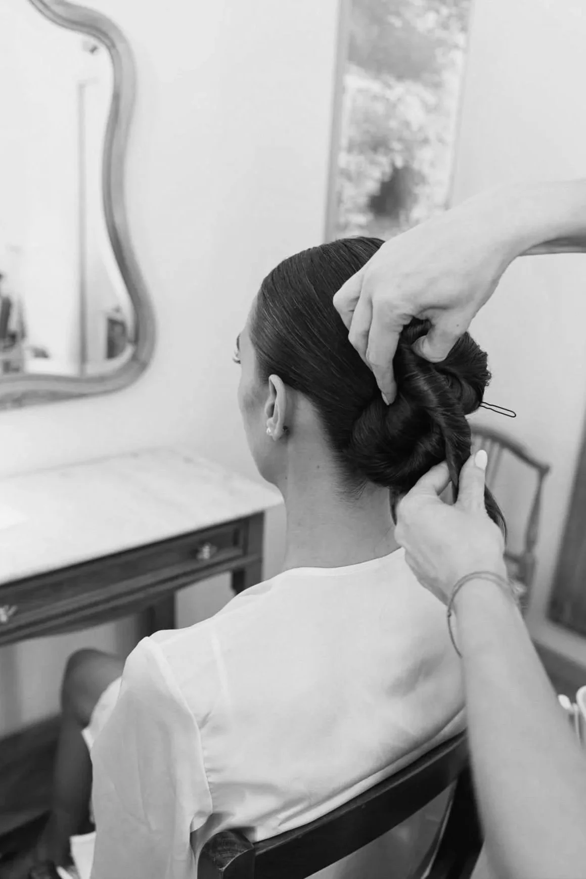 Une femme est assise dans une chaise, avec les mains d'une coiffeuse entrain de construire un chignon de mariage.