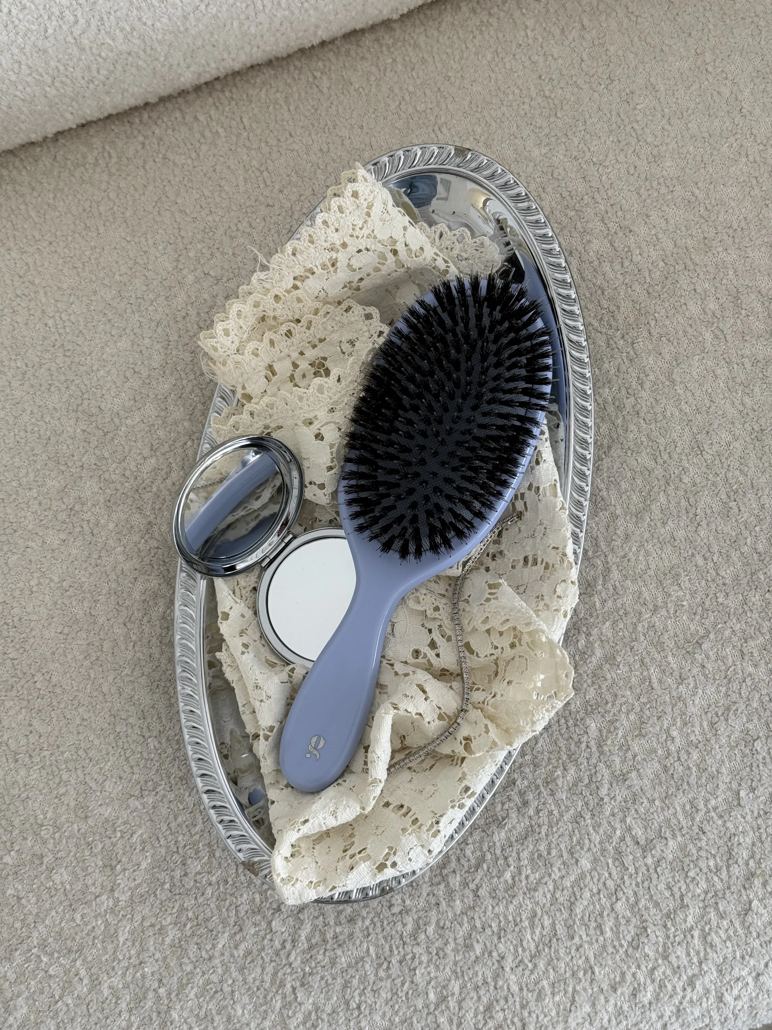 Silver tray with lace cloth, a light purple hairbrush with black bristles, a small round mirror, and a round compact mirror, all placed on a beige carpeted surface.