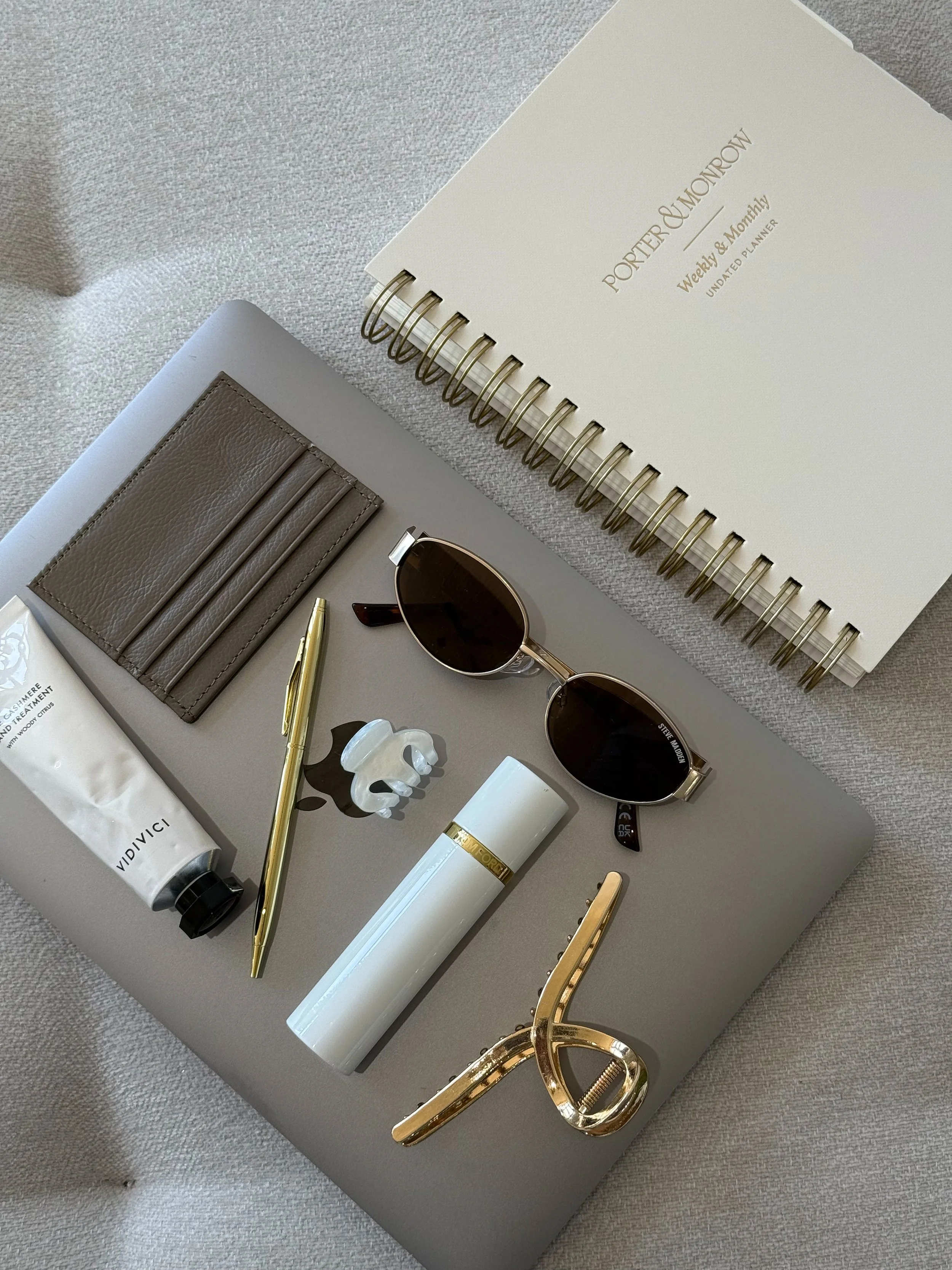 Flat lay of a laptop with sunglasses, a wallet, a tube of hand cream, a gold pen, a white spray bottle, a gold hair clip, a white hair clip, and a white undo list notebook.