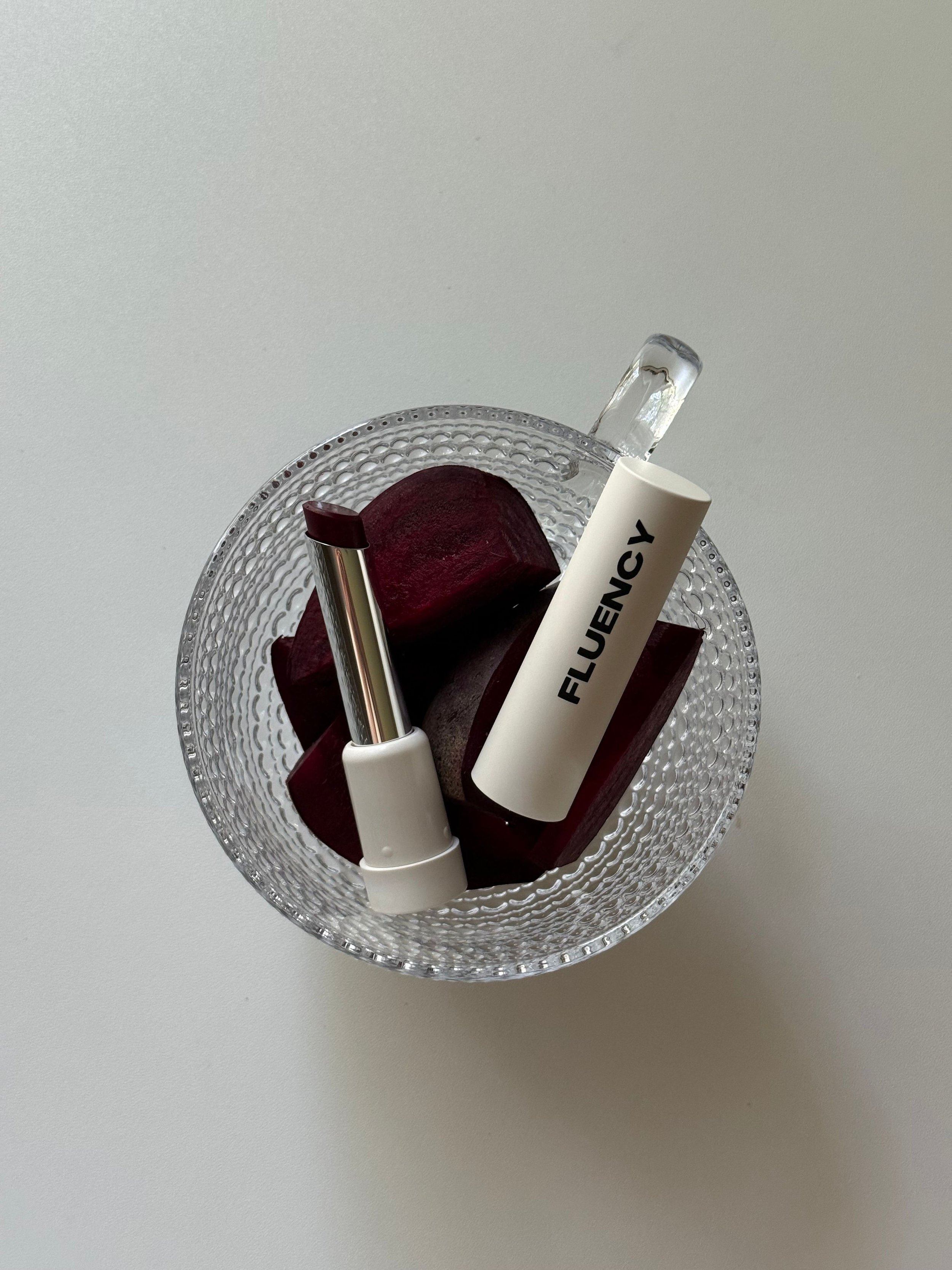 A glass bowl containing two lipstick tubes and several slices of beets on a white surface.
