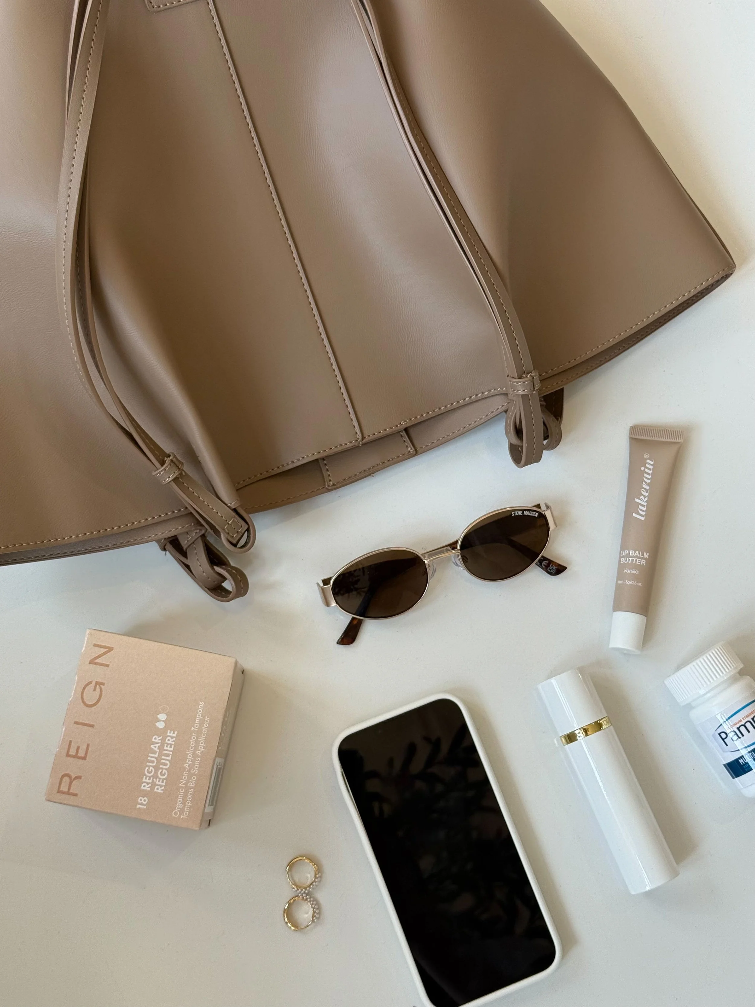 A beige handbag, sunglasses, a tube of lip balm, a box of tampons, a smartphone, a bottle of vitamins, a small white container, and a pair of earrings on a white surface.