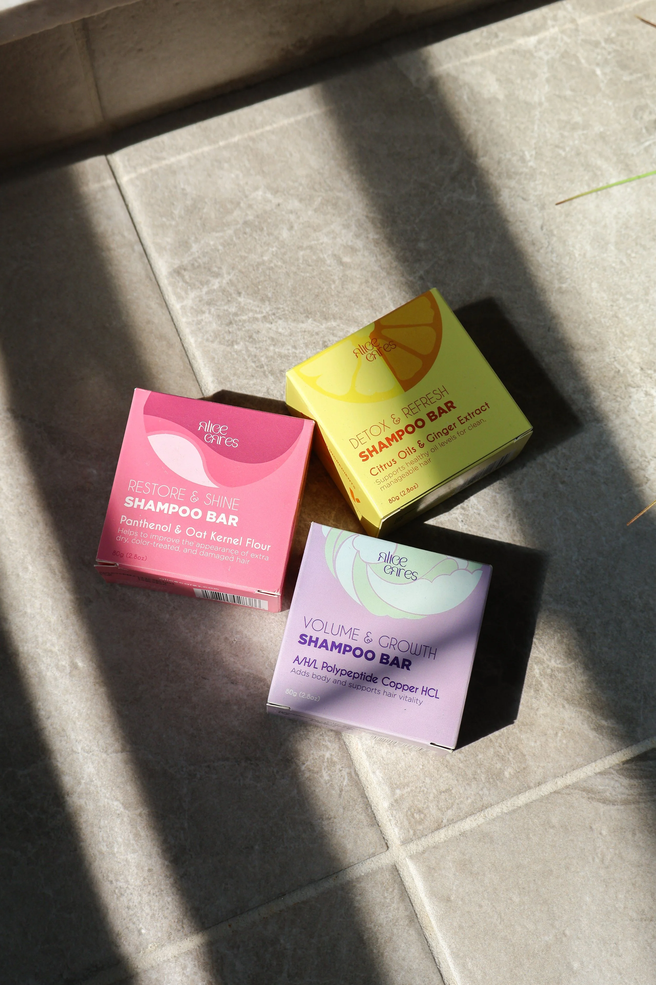 Three boxes of Alice Cares shampoo bars in pink, yellow, and purple, placed on a tiled floor with sunlight casting shadows.