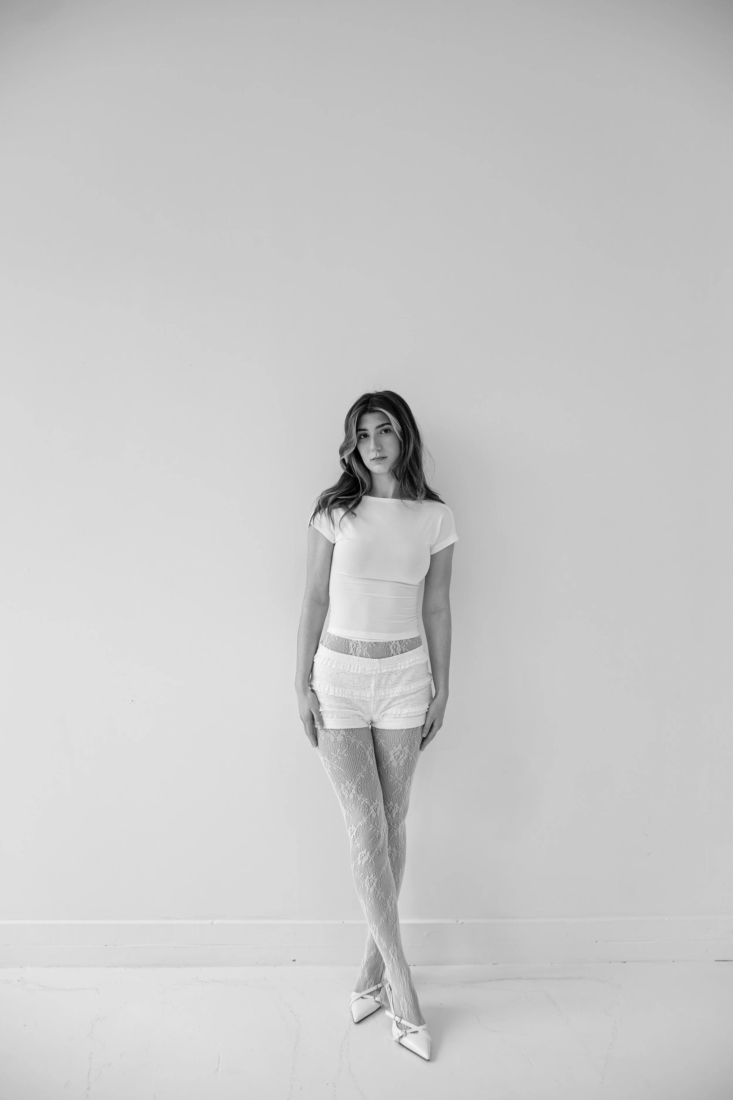 A young woman with wavy hair, dressed in a white t-shirt, shorts, lace tights, and high heels, standing against a plain, white wall in black and white.