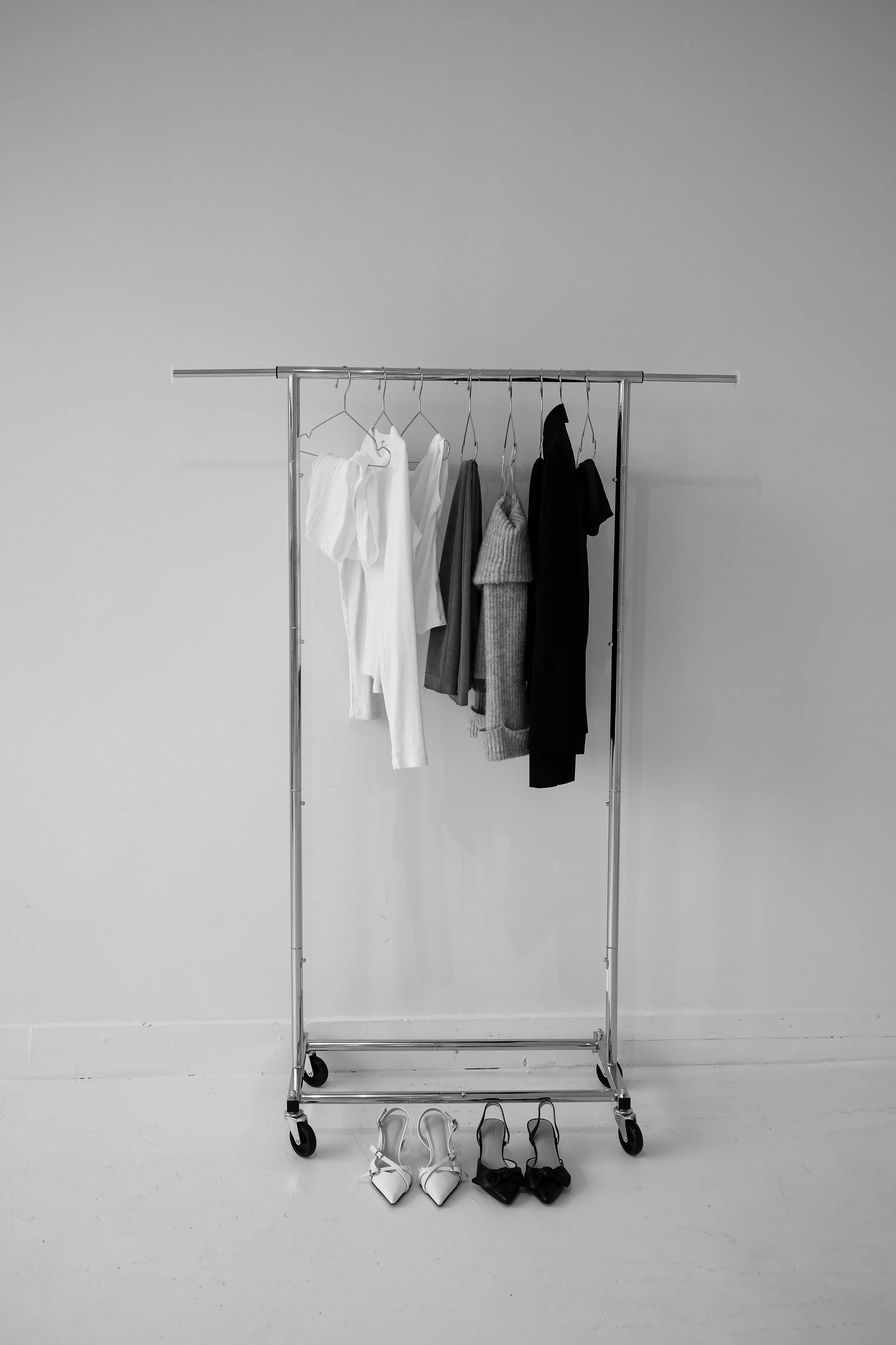 A metal clothing rack with five hanging garments and two pairs of high-heeled shoes placed on the floor in front of it, set against a blank wall.