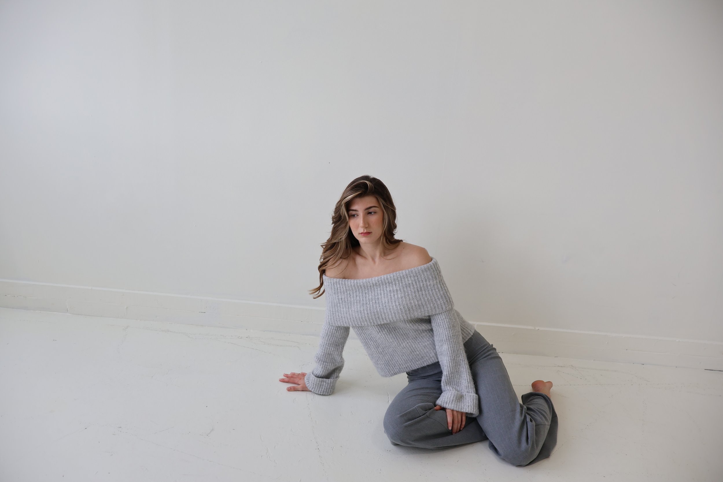A young woman with wavy brown hair sitting on the floor against a plain white wall, wearing a gray off-the-shoulder sweater and gray pants.