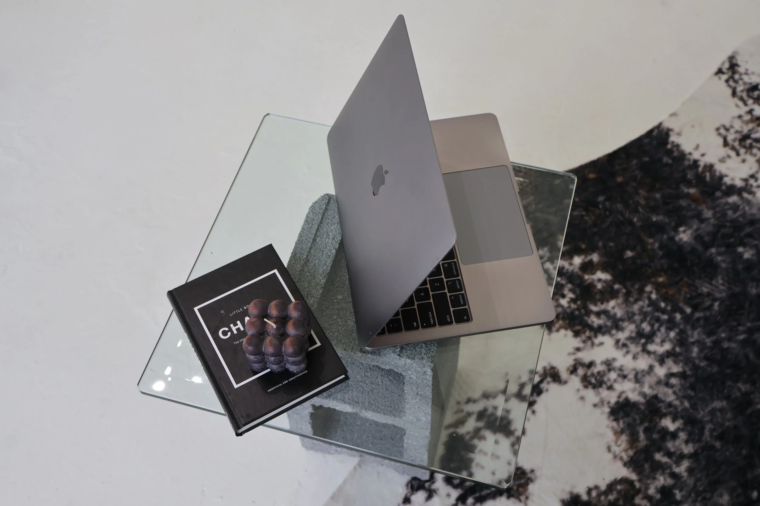 A silver MacBook laptop partially opened on a glass table with a black book and a decorative object on top, set against a background of a white floor and a textured black and white wall.