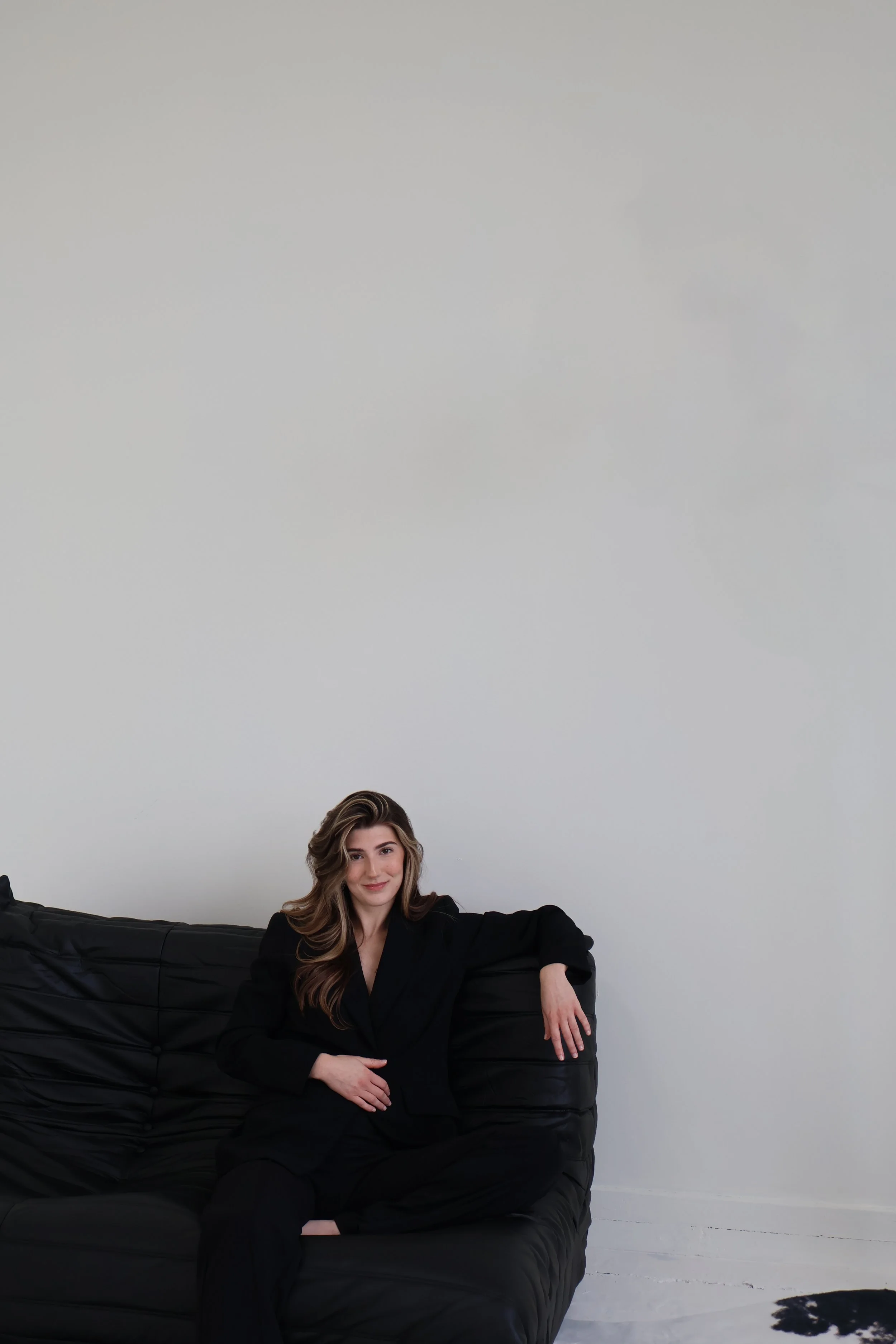 A woman with wavy hair sitting on a black leather couch in front of a plain white wall, wearing a black suit and smiling.