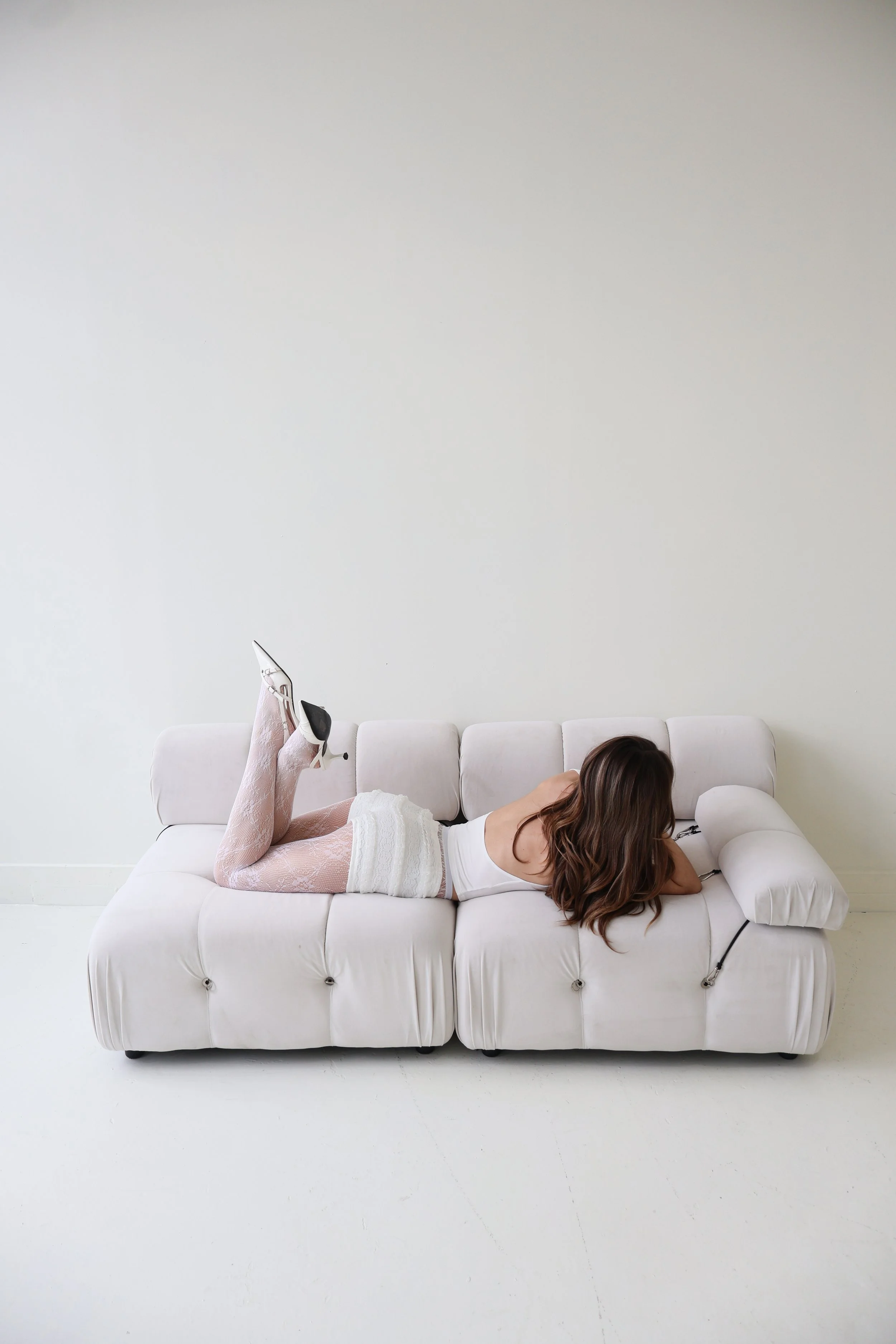 A woman lying on her stomach on a white sofa, wearing white lace stockings, a white top, and high heels, with her head resting on her arms.