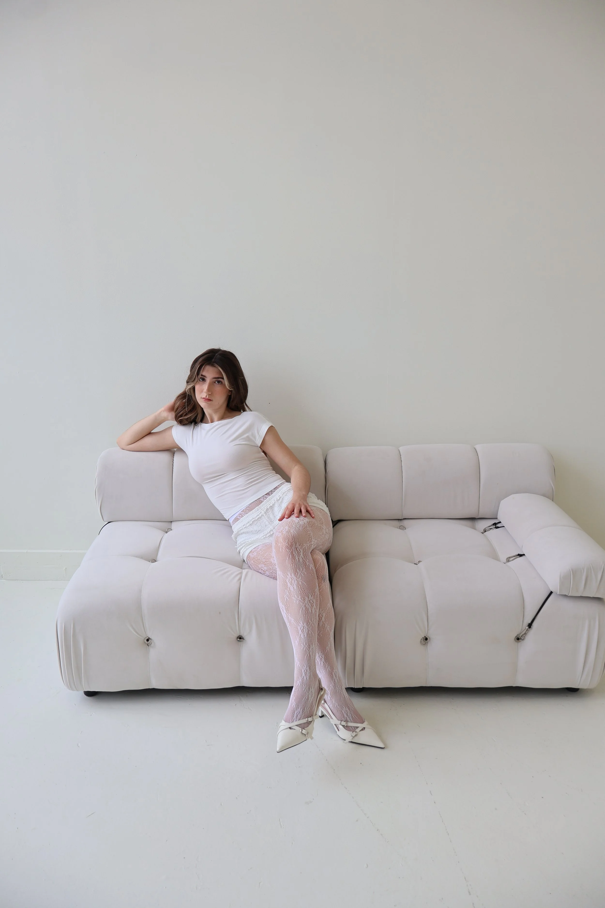 Young woman in white attire with patterned tights and high heels sitting on white sofa in minimal room.