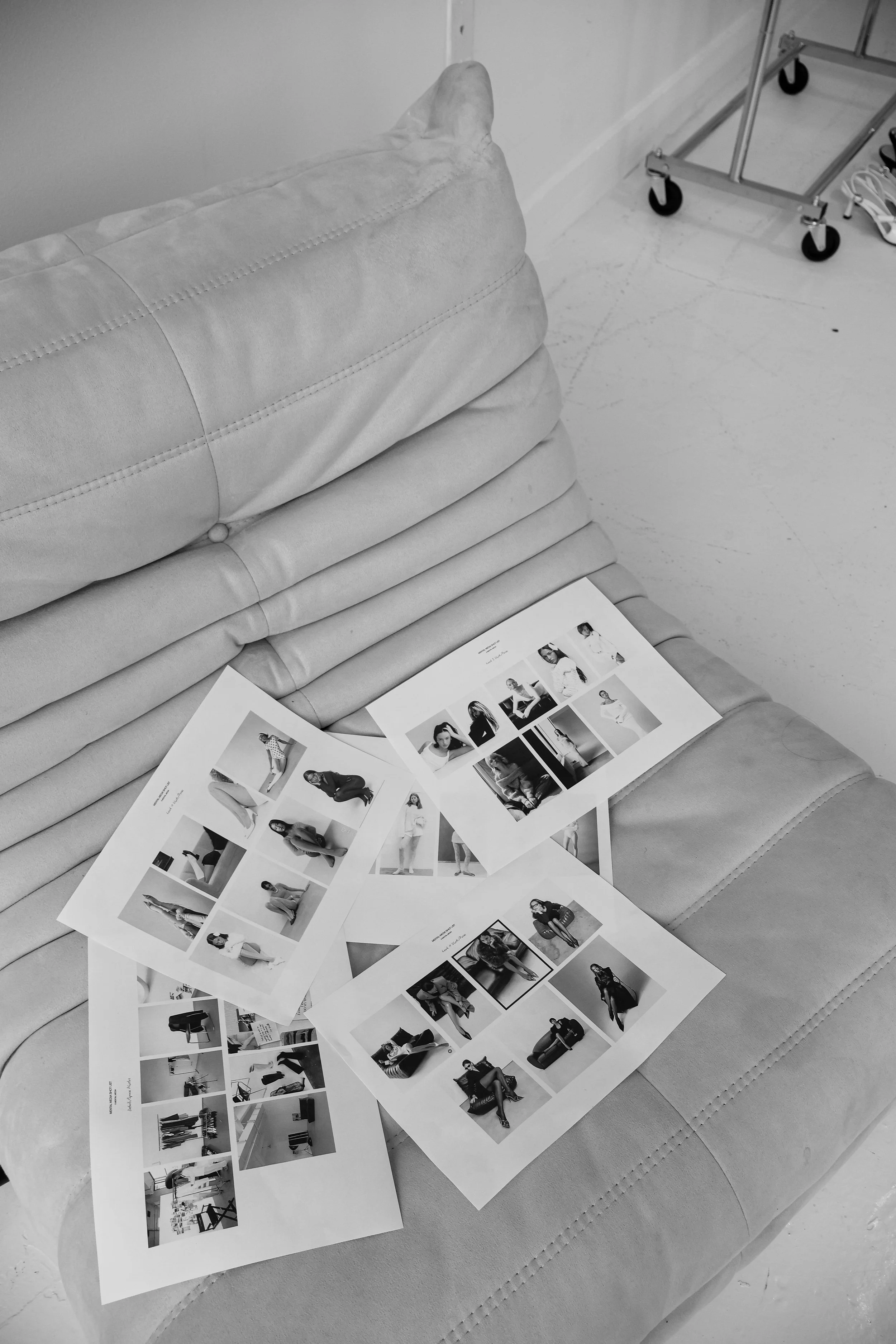 Black and white photograph of printed photos of models on a beige, cushioned sofa. The photos are laid out in a grid pattern, showcasing various poses and outfits.