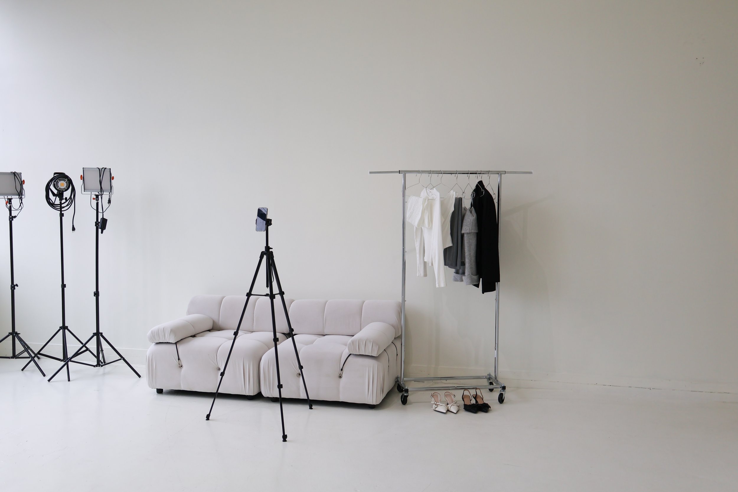 A minimalist photo studio setup with three photography lights, a camera on a tripod, a white sofa, a clothing rack with five garments, and three pairs of shoes on a white floor.