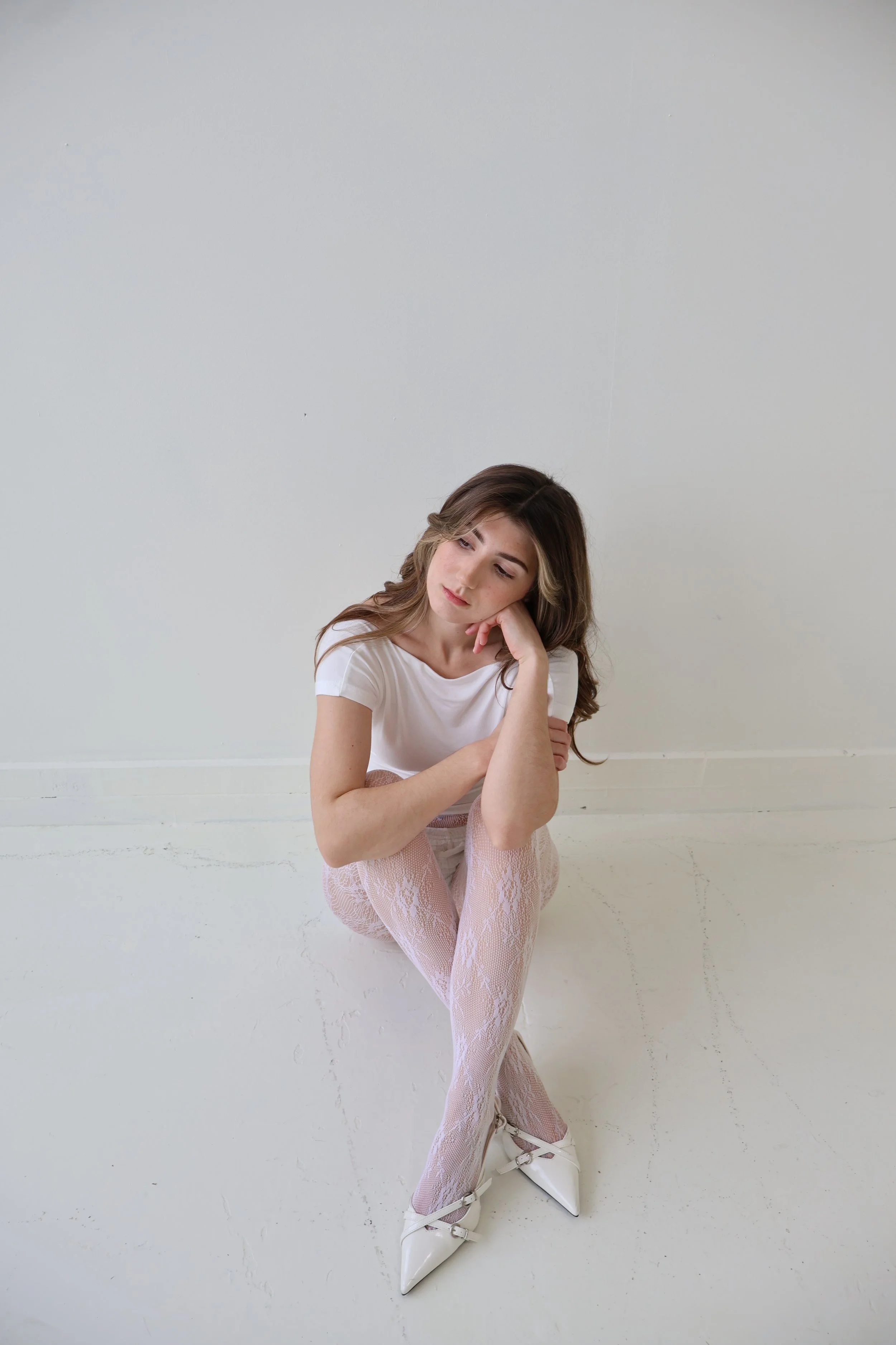 Young woman sitting on the floor, resting her head on her hand, wearing a white t-shirt, pink lace tights, and white high-heeled shoes, against a plain white wall.