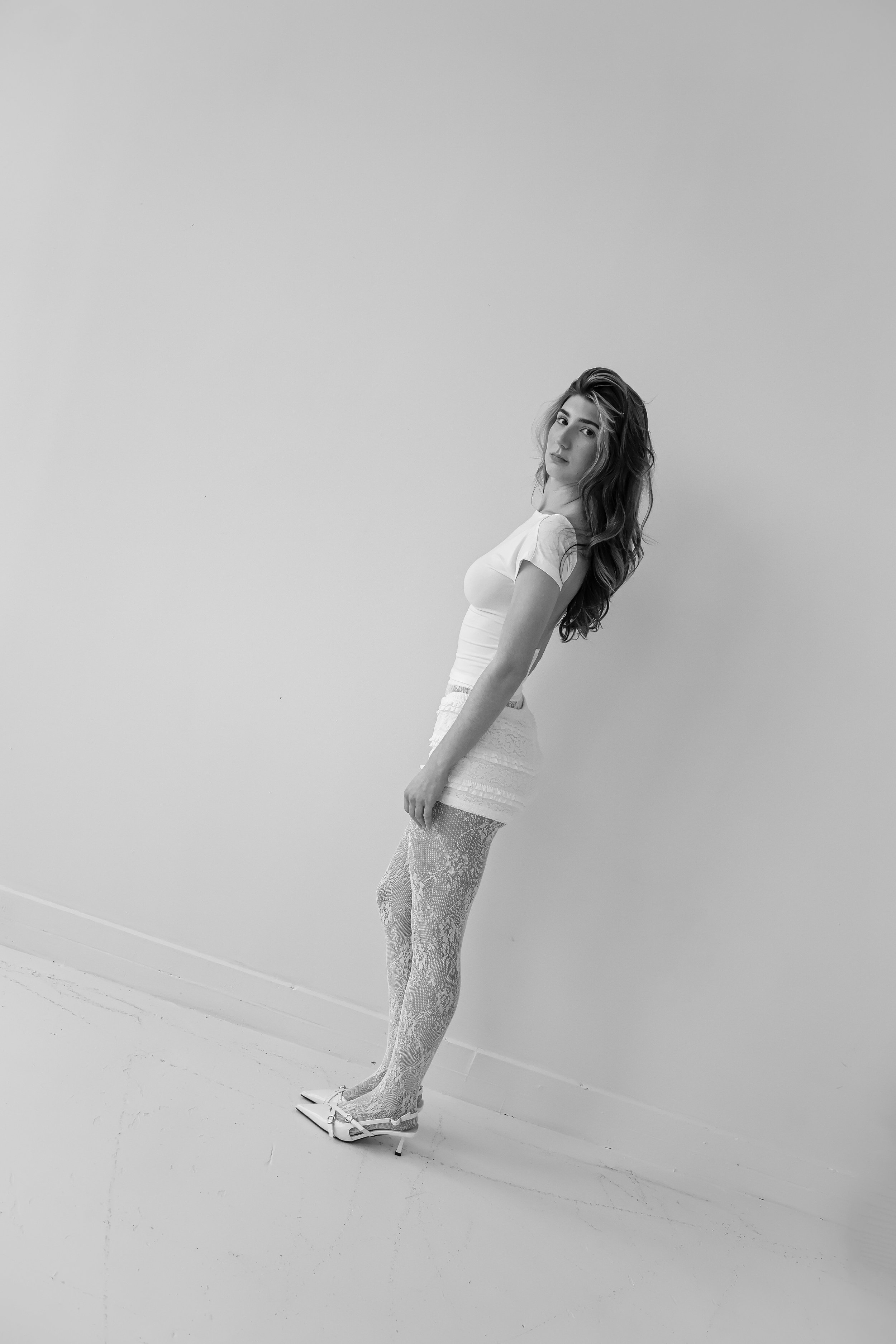 A woman with long hair in a white t-shirt and lace tights standing against a plain wall, looking at the camera.