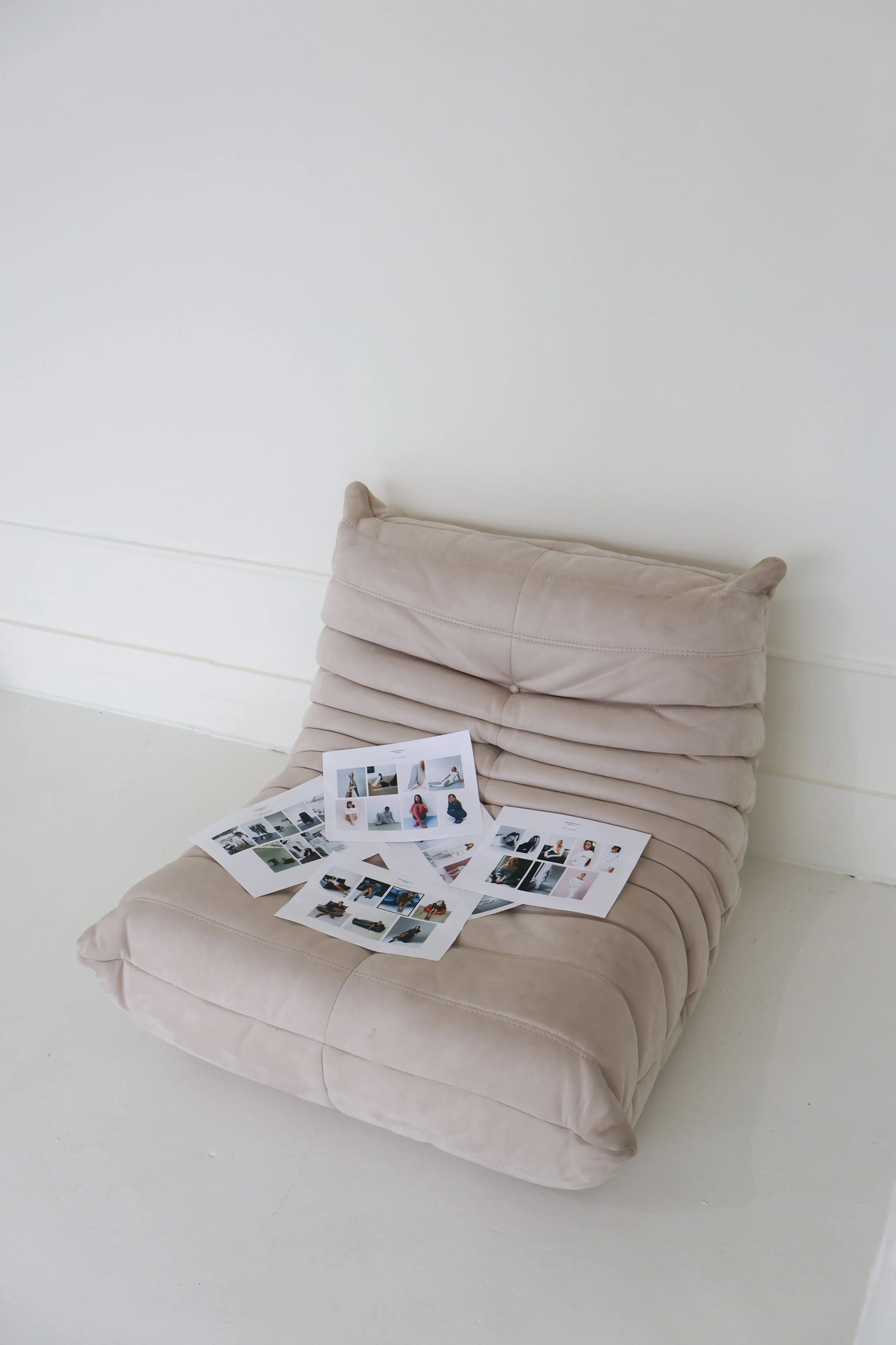 A beige cushioned chair with several printed photographs spread out on it, placed against a plain white wall.