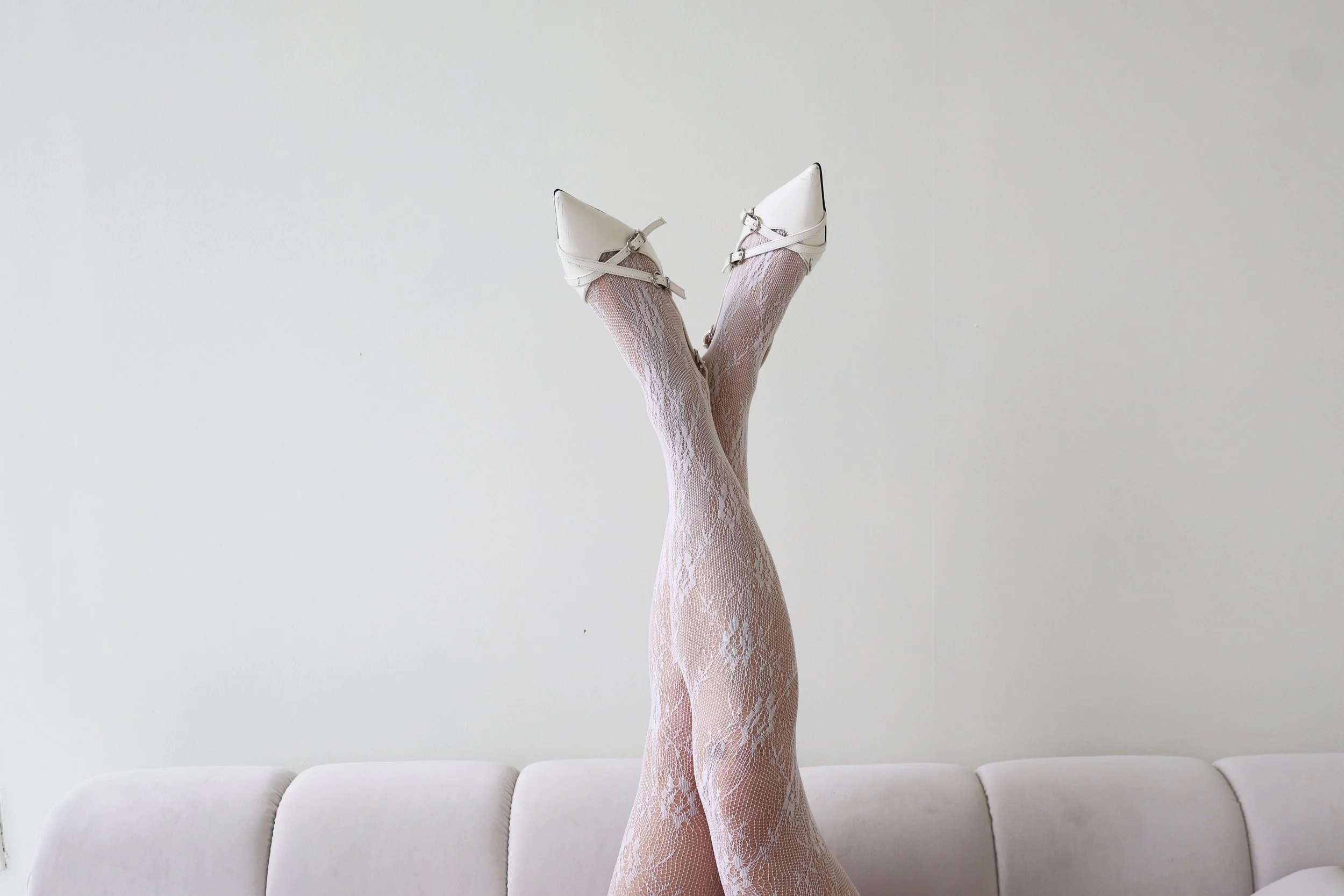 Person lying on a sofa with legs raised and crossed, wearing white lace stockings and white high-heeled shoes.