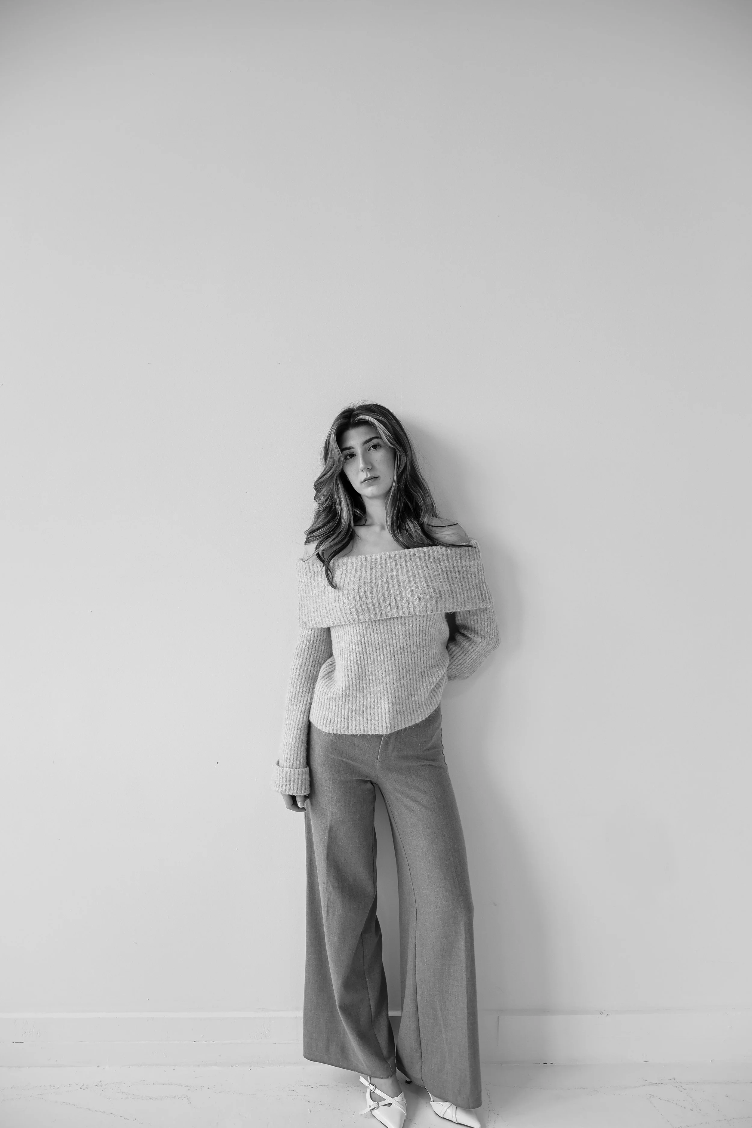 Black and white photo of a woman with wavy hair standing against a plain wall, wearing a cropped off-the-shoulder sweater and wide-legged pants.