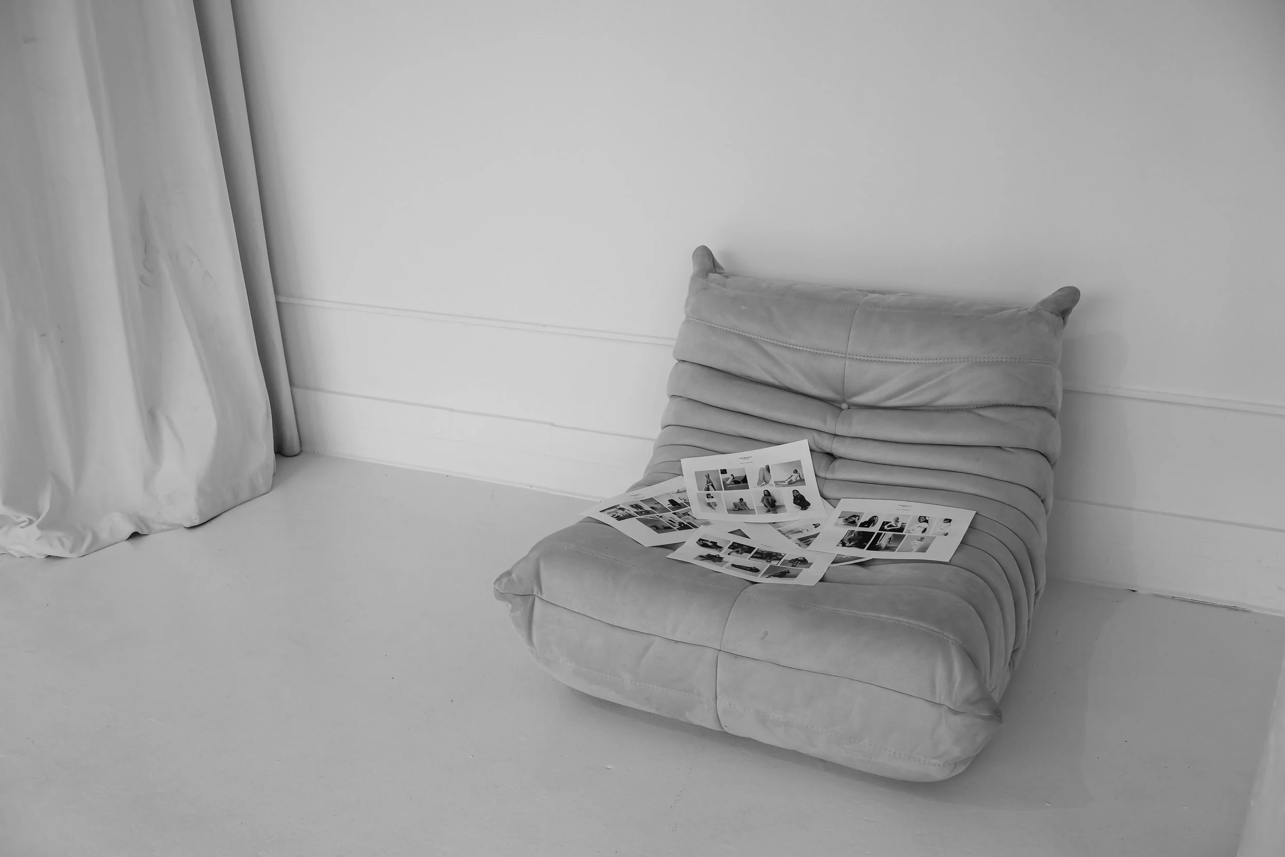 A portable stuffed chair with horizontal stitching, placed on a light-colored smooth floor, with several printed photos or posters on it. To the left, white curtains are partially visible, hanging beside a white wall.