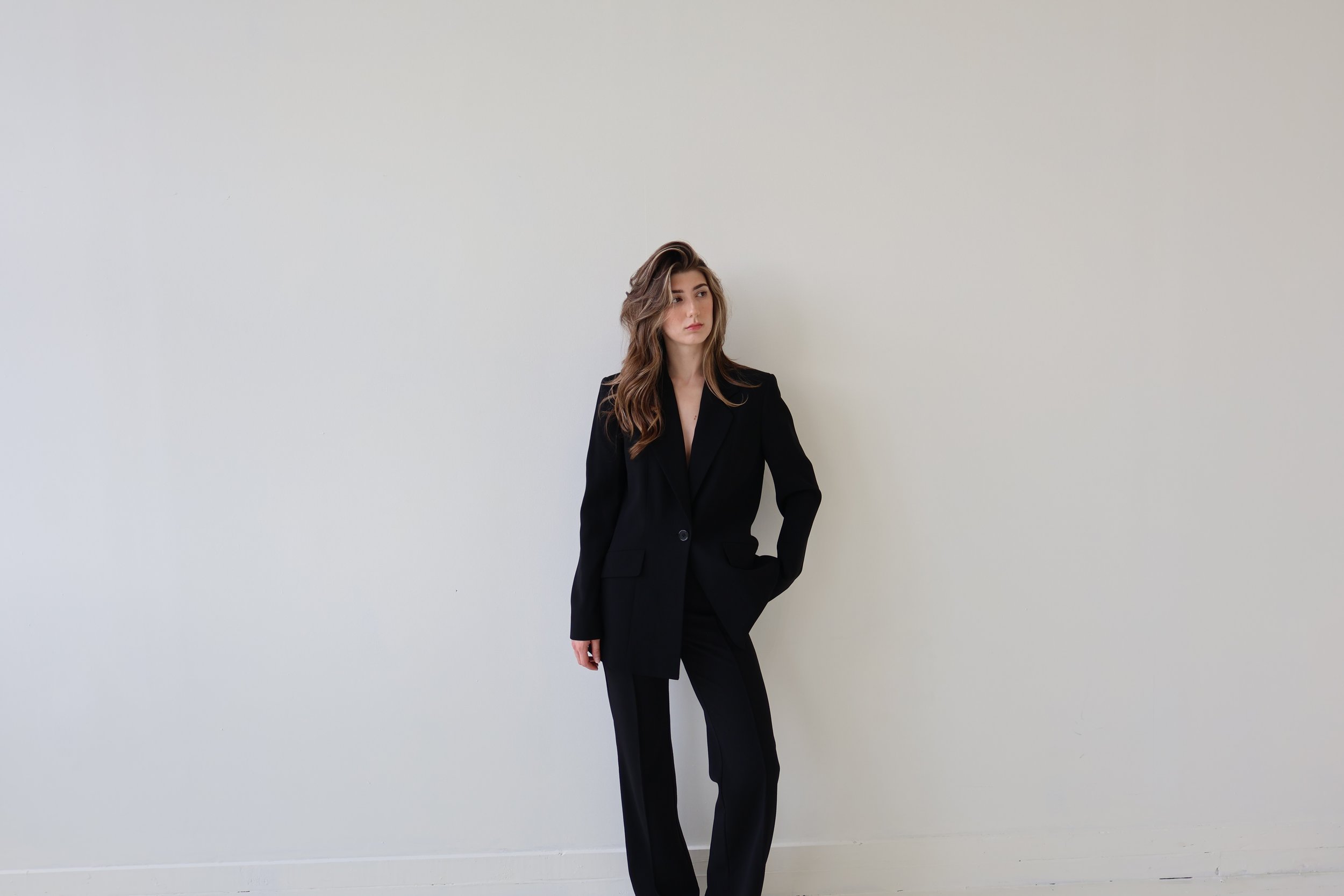 Young woman in black suit standing against a plain white wall.