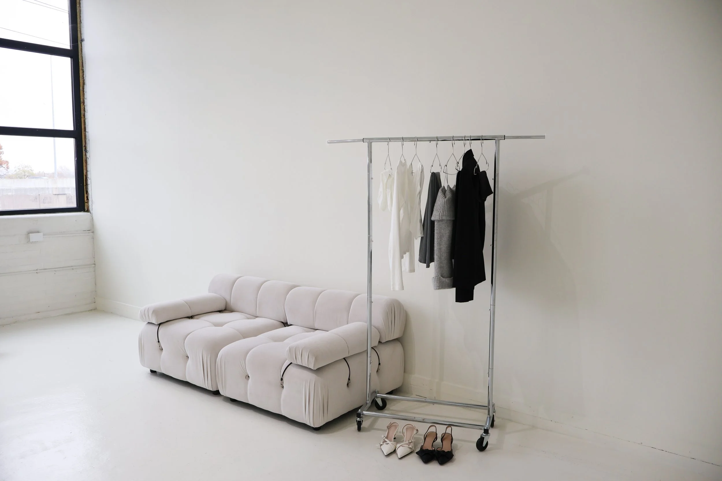 A clothing rack with white, gray, and black clothes, and two pairs of shoes, one white and one black, are in front of a white sofa in a bright, minimalist room with a large window.