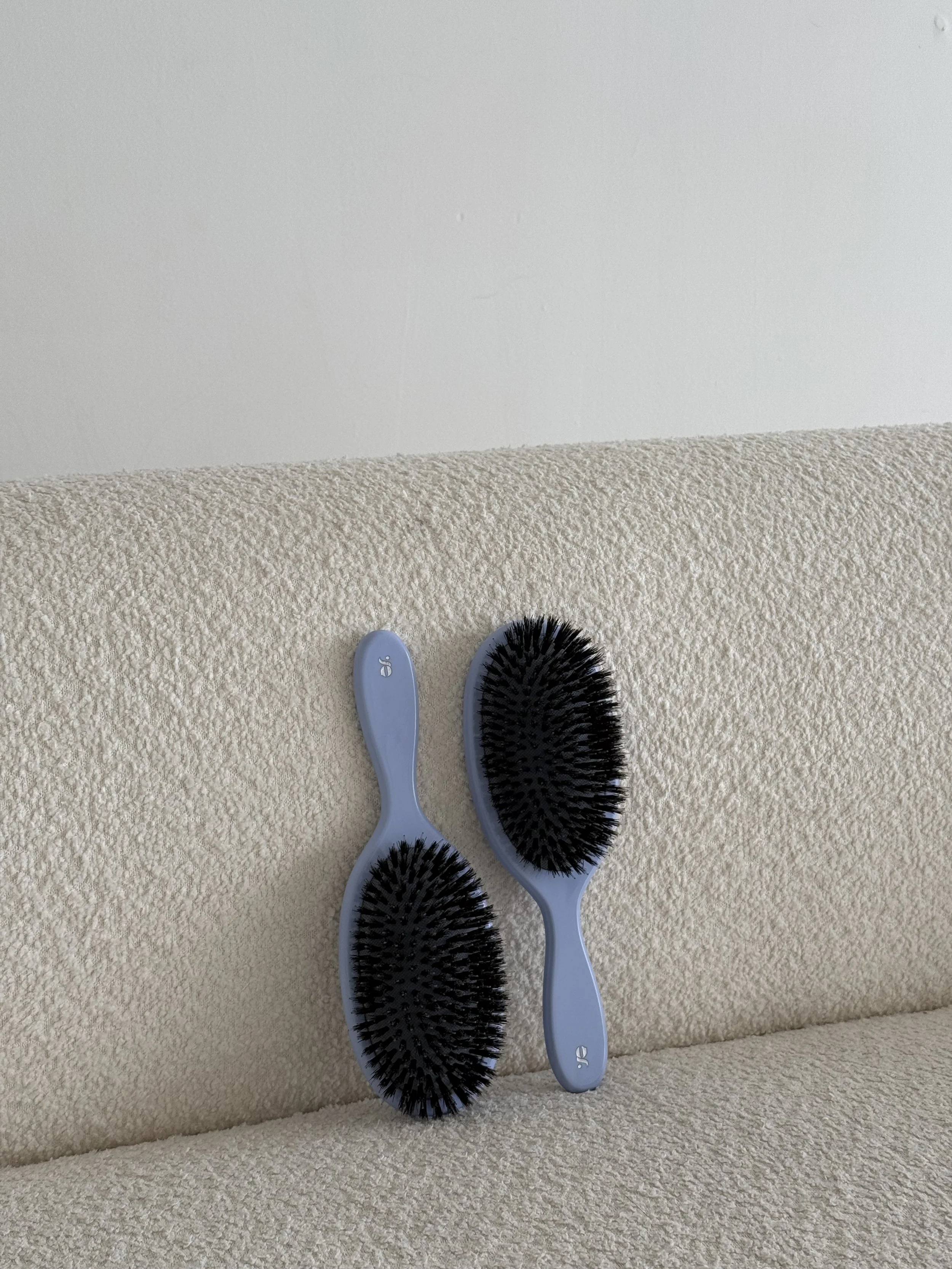 Two hairbrushes with black bristles and light blue purple handles, resting on a beige textured sofa, against a white wall background.