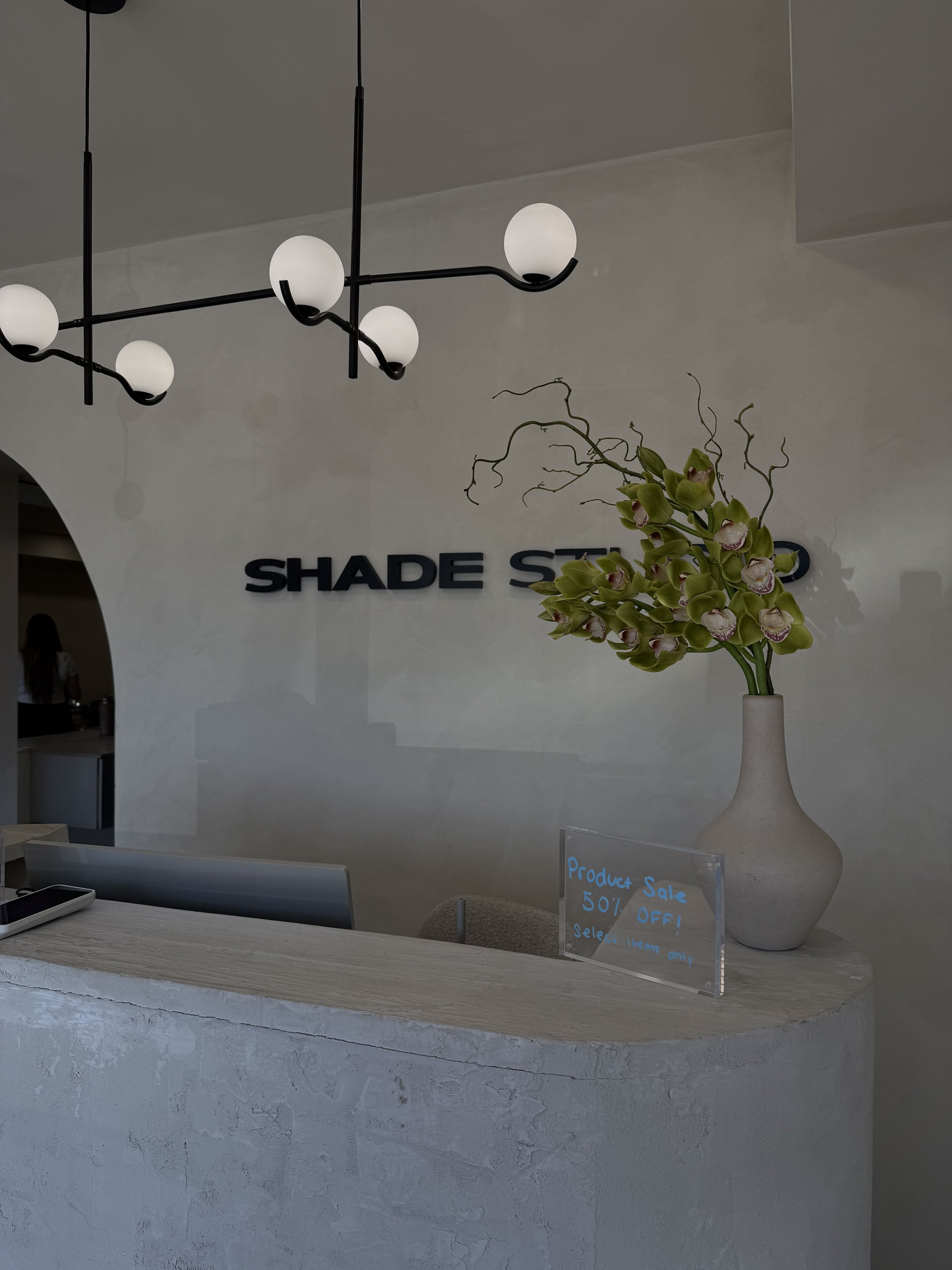 A reception desk with a large white vase holding green orchids, a sign advertising a 50% off product sale, and a modern chandelier hanging above. The background features a wall with the partial name "SHADE" on it.