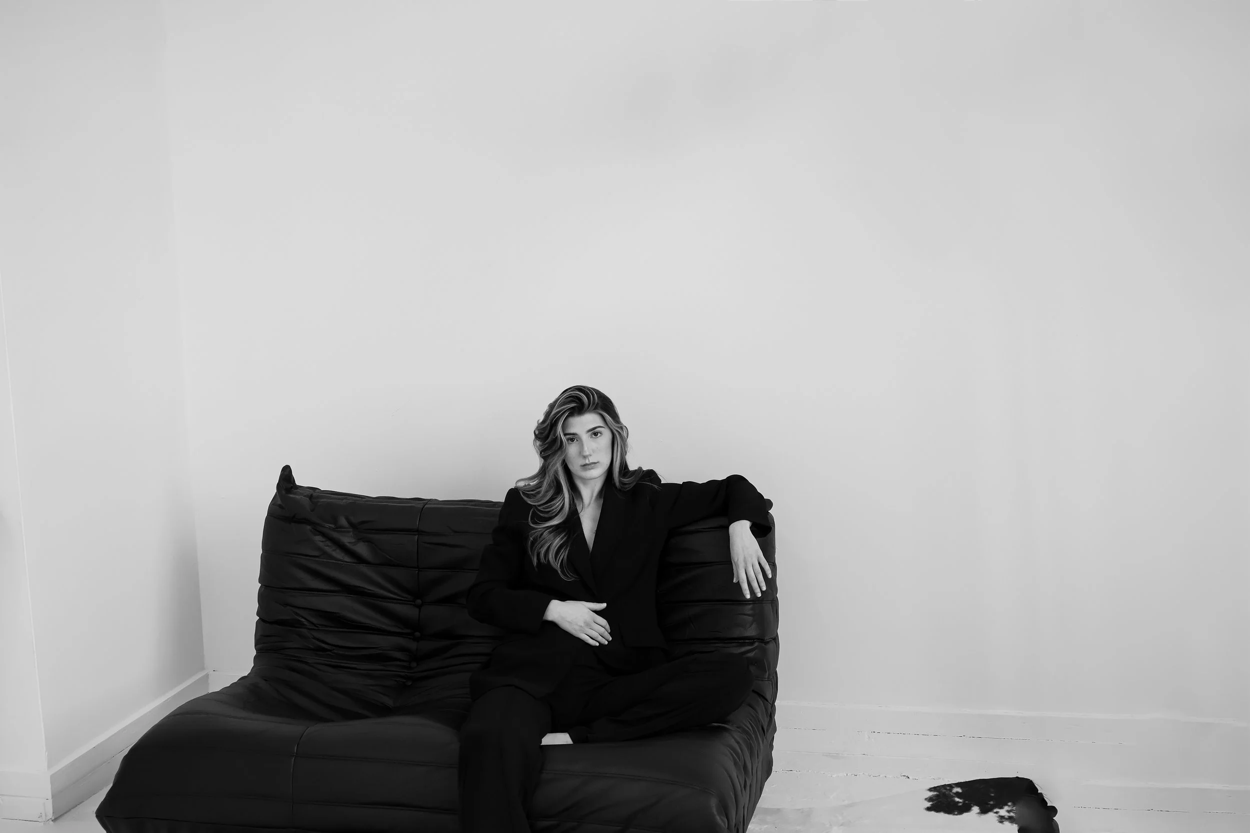 Black and white photo of a woman sitting on a modern, black, leather sofa in a minimalistic room with white walls and wooden floor. She has long, wavy hair and is dressed in a black suit, looking directly at the camera.