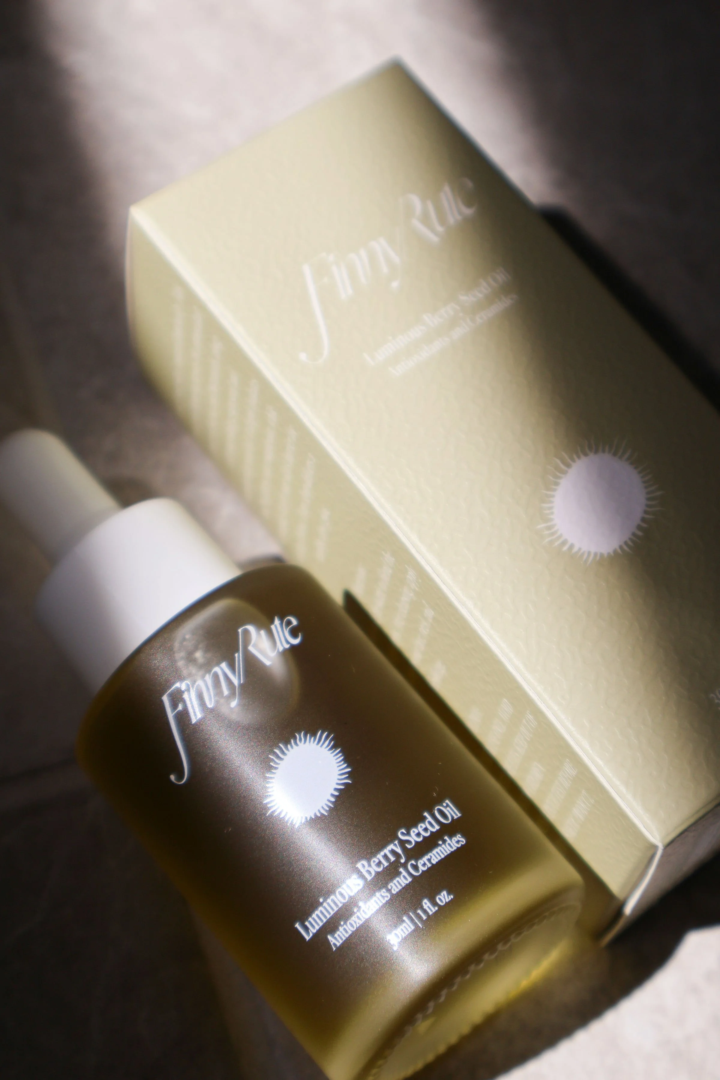 A bottle of Finny Rute luminous berry seed oil with a white cap and a beige box in the background, on a gray surface.