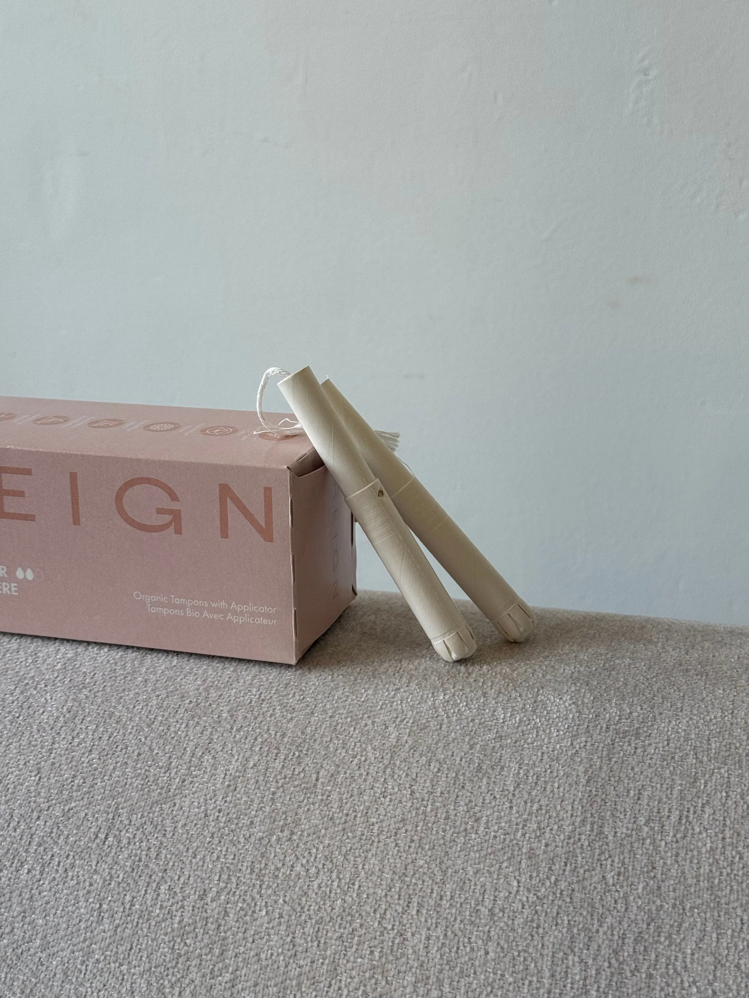 A pink box labeled 'Organic Tampons with Applicator' with two beige tampons leaning against it on a gray surface, against a white wall background.