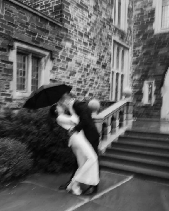 A black and white photo of a couple embracing on a rainy day outside a brick building, with the man holding an umbrella.