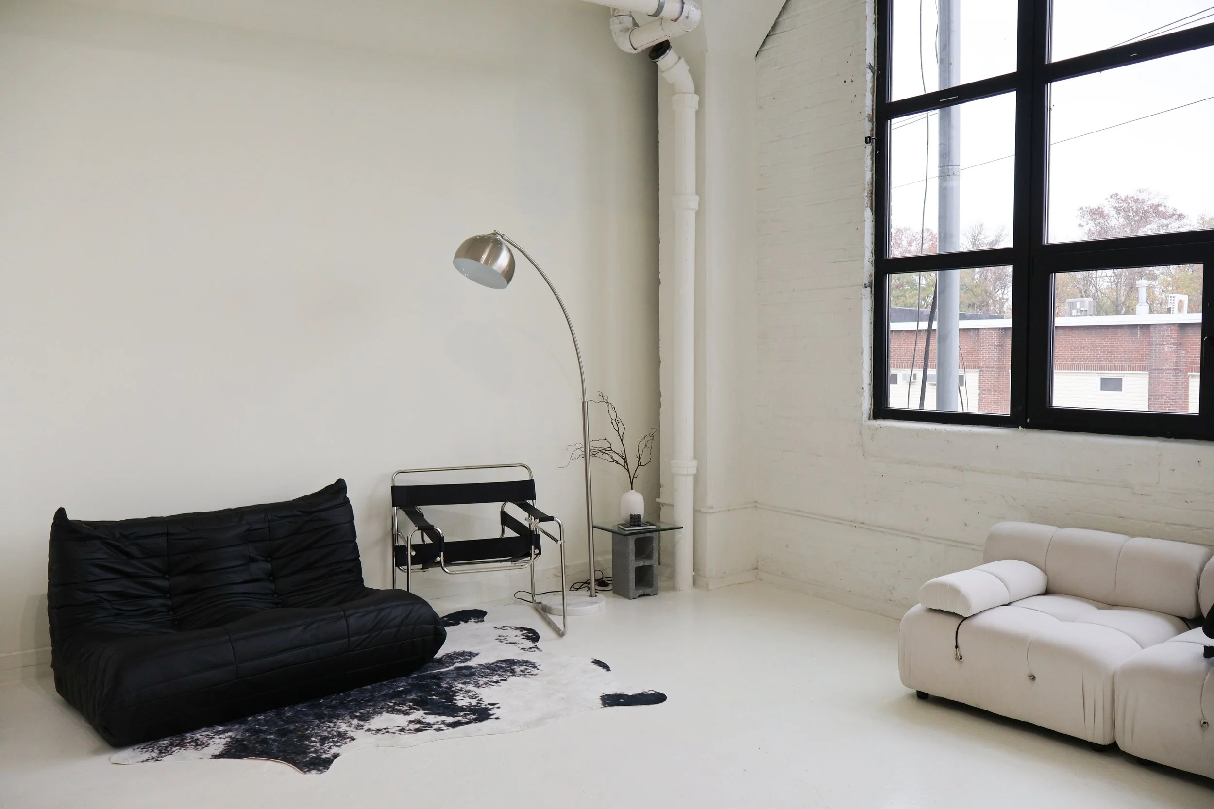 Minimalist living room with black and white sofas, a floor lamp, a small side table with a vase, and large black-framed windows showing an urban exterior.