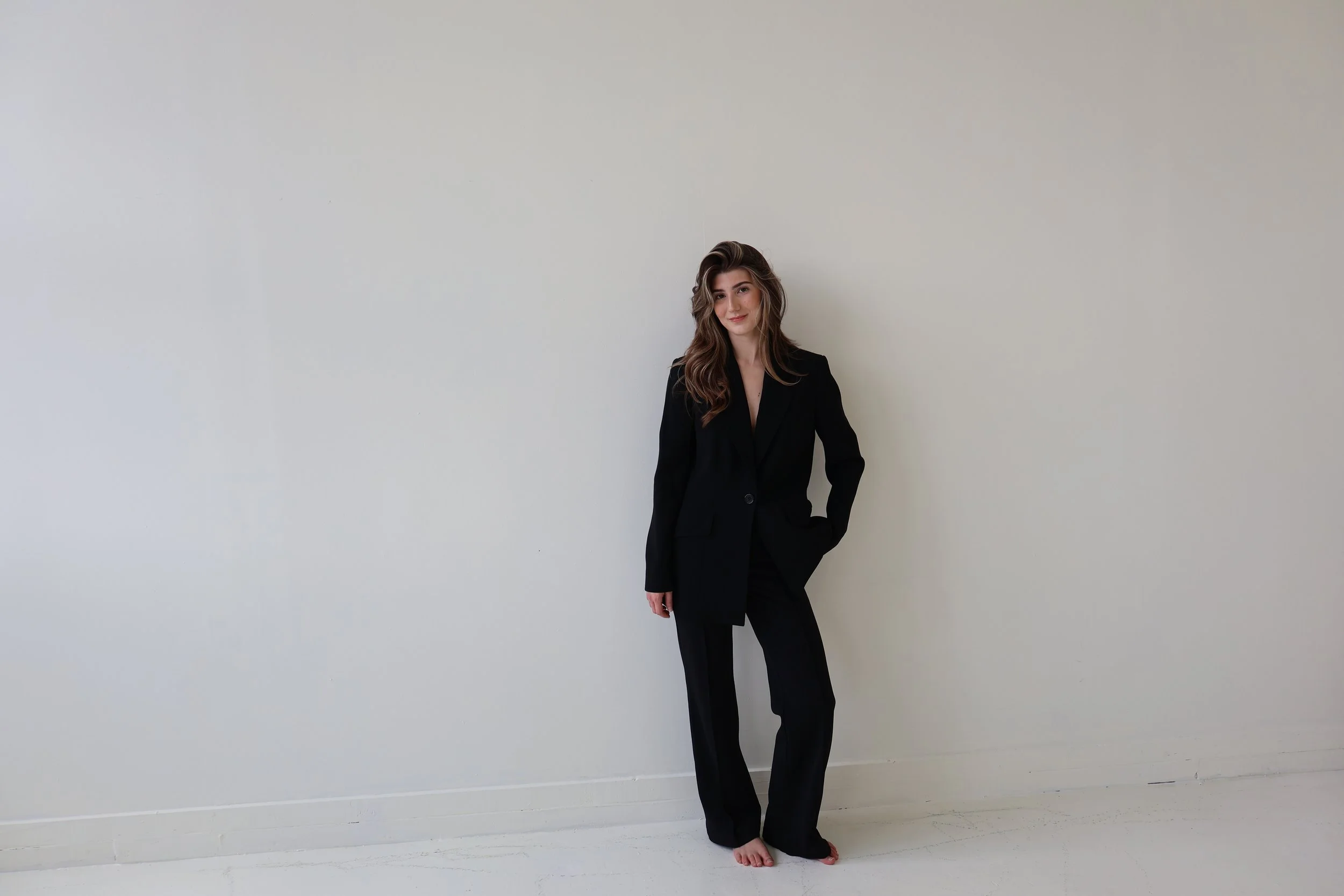 A young woman with long wavy brown hair wearing a black blazer and black pants, barefoot, standing against a plain white wall.